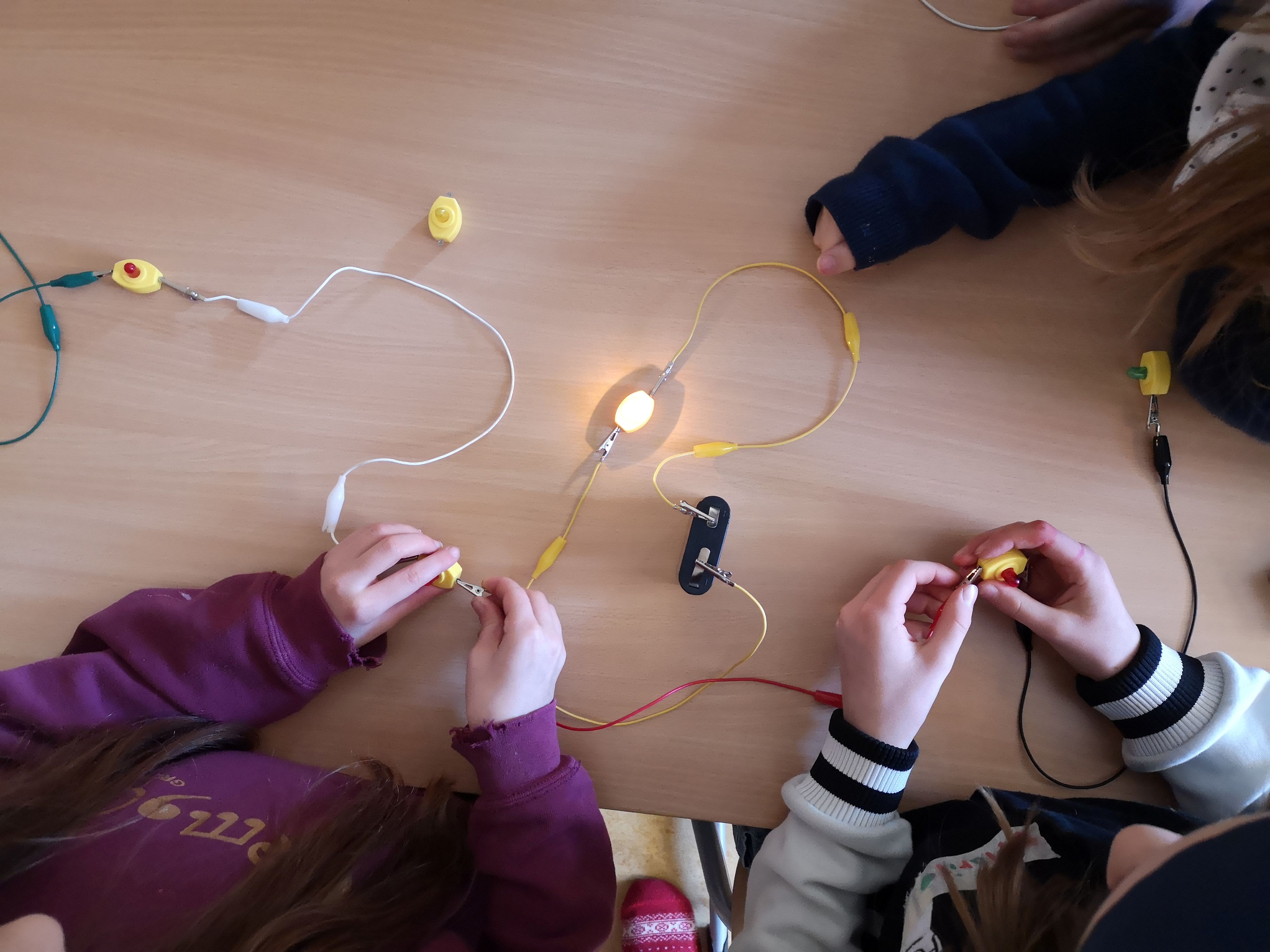 Mehrere Kinder experimentieren mit einem einfachen Stromkreis auf einem Tisch. Kabel, Klemmen und Schalter verbinden eine Batterie mit einer leuchtenden Glühbirne, während die Kinder die Verbindungen testen.