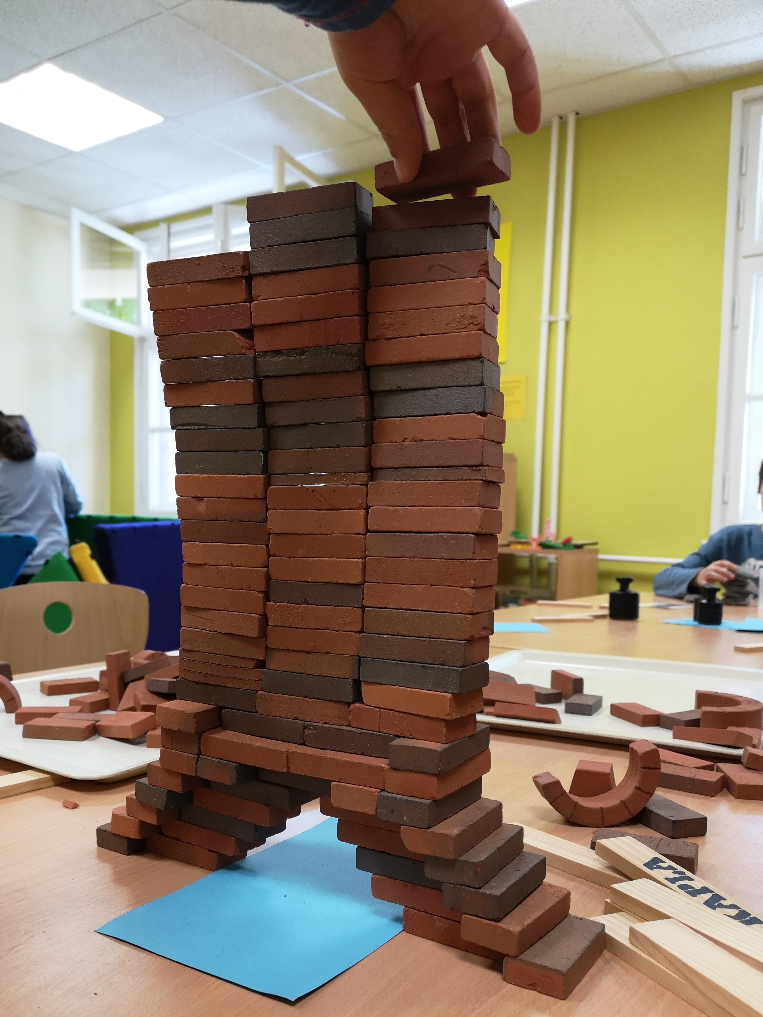 Eine Hand setzt einen rechteckigen Baustein auf die Spitze eines hohen Turms aus kleinen rotbraunen und dunklen Bausteinen. Die Konstruktion steht auf einem Tisch und ruht auf zwei gestuften Pfeilerfüßen, während weitere Bausteine und Baumaterialien im Hintergrund liegen.