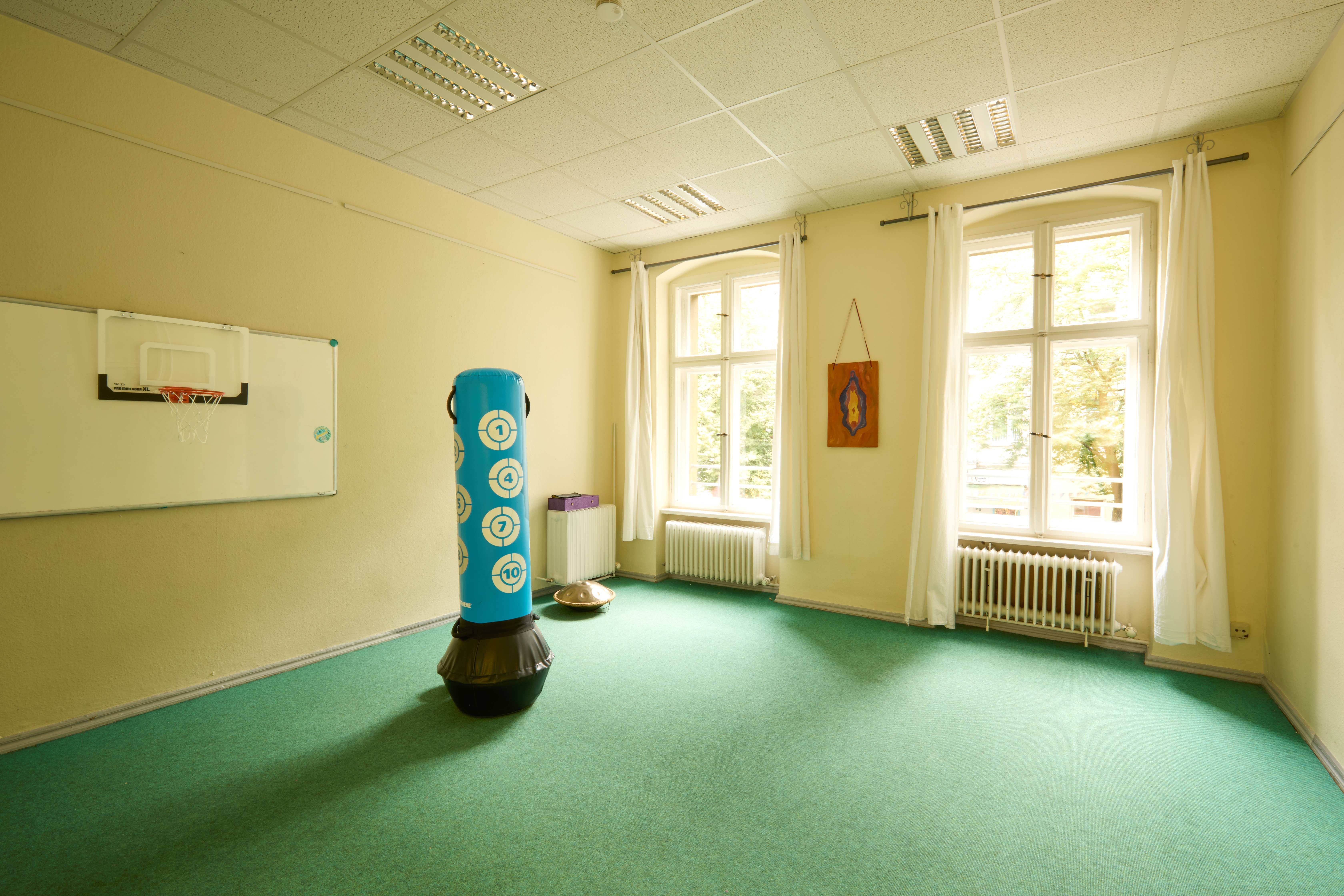 Heller Sportraum mit grünem Teppich, zwei hohen Fenstern, Basketballkorb an der Wand und blauem Boxsack in der Mitte.