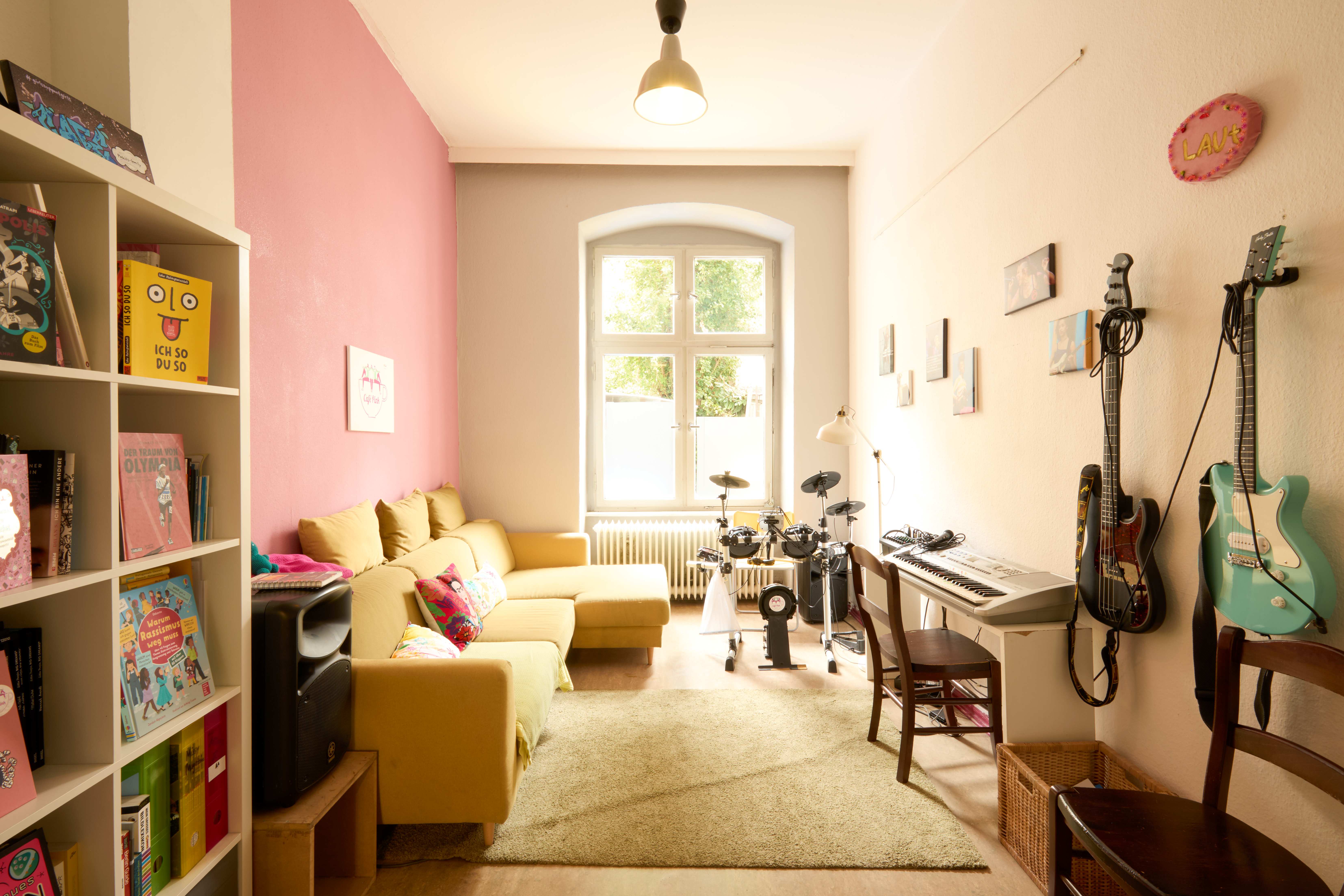 Gemütlicher Musik- und Leseraum mit gelbem Ecksofa, Regal mit Büchern, E‑Drumset, Keyboard und an der Wand hängenden E‑Gitarren.