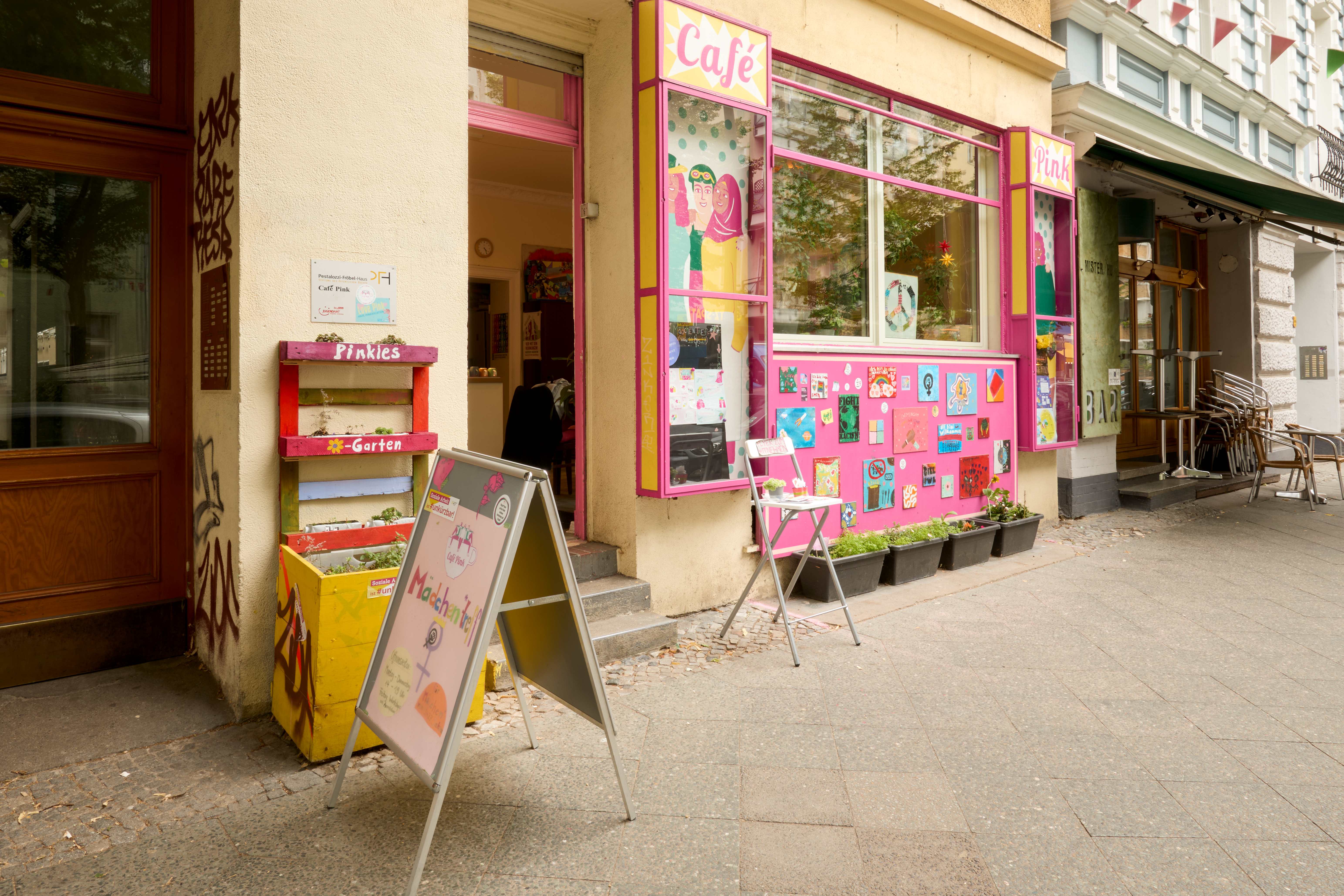 Außenansicht eines Gebäudes mit dem farbenfrohen Eingang des Café Pink, großen Fenstern und Pflanzkästen; am Eingang stehen Tische und Stühle.