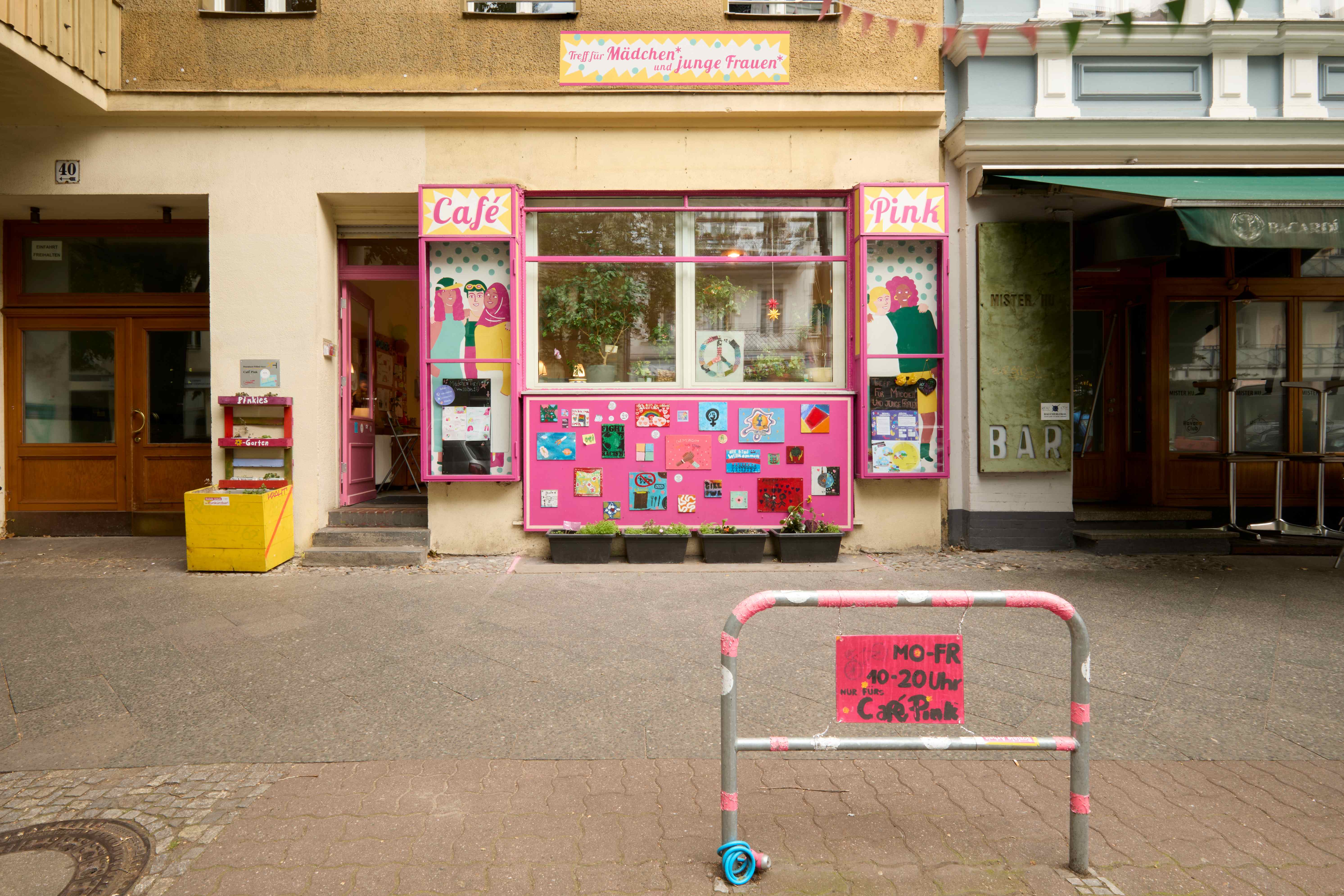 Gebäudefassade mit pink umrahmten Fenstern und dem Schild „Treff für Mädchen* und junge Frauen*“; darunter ist der Eingang zum Café Pink.