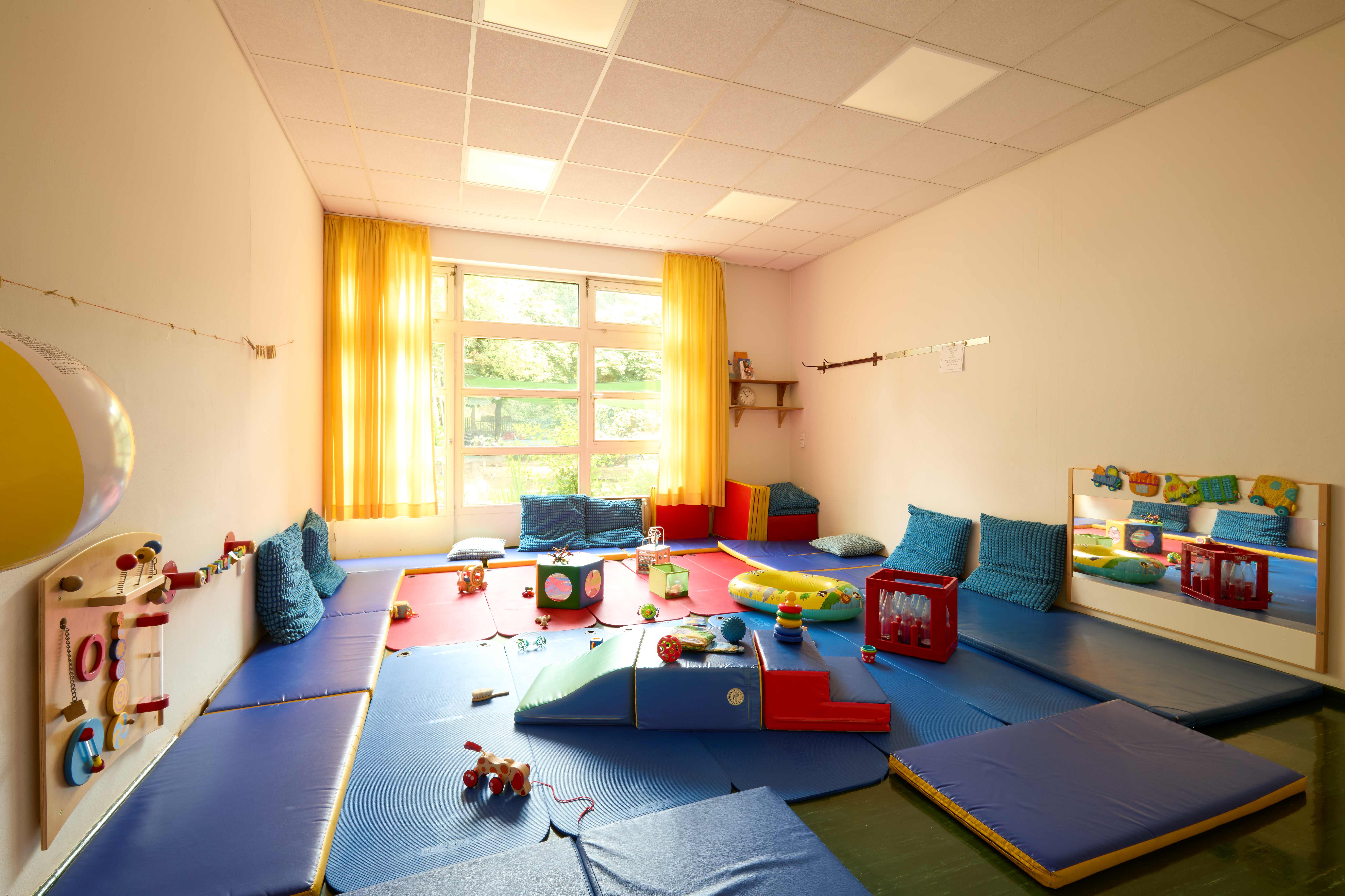 Bewegungsraum mit blau ausgelegten Matten, gelben Vorhängen und vielfältigen Motorikmaterialien sowie Spielzeugen für Kleinkinder.