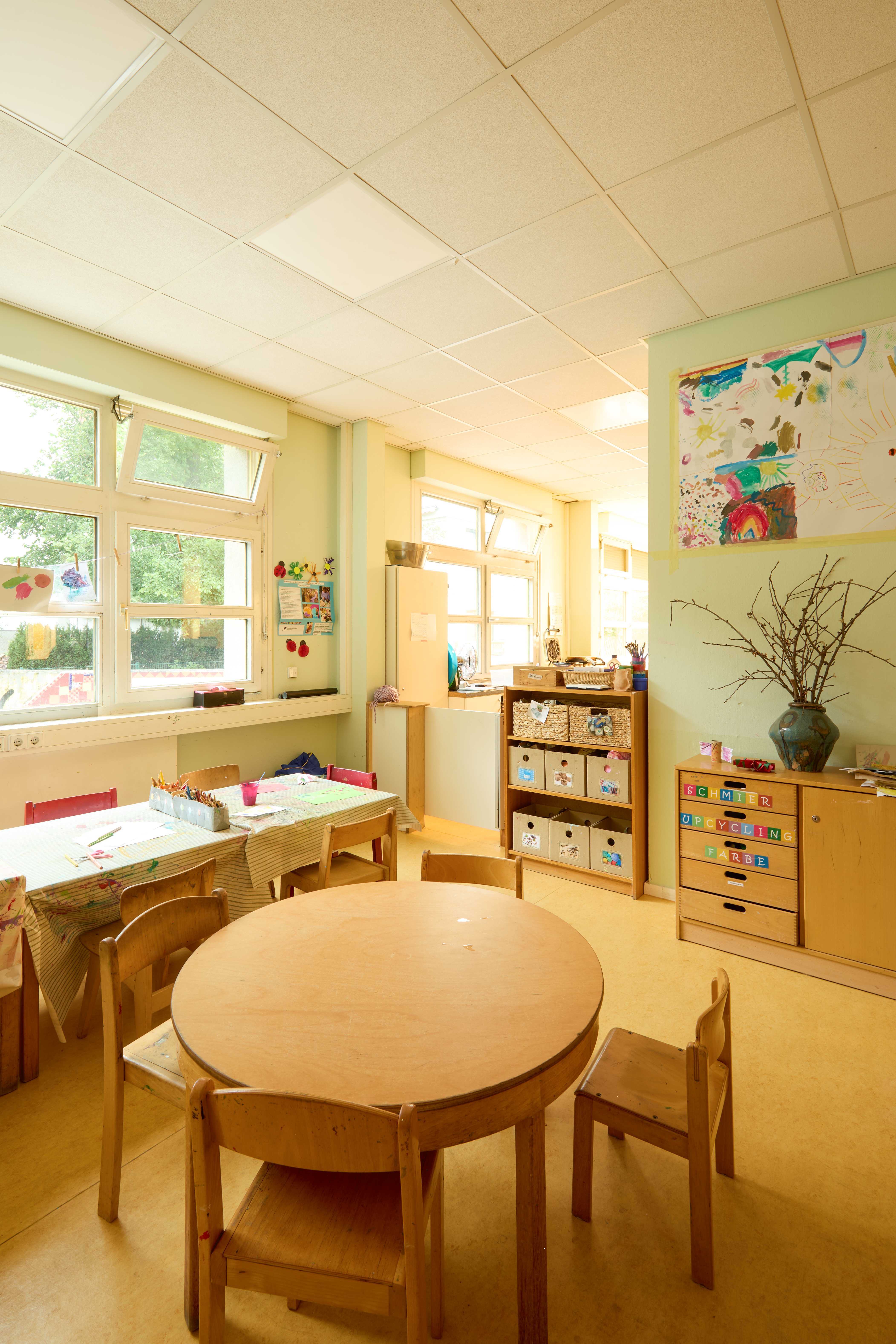 Heller Kreativ‑ und Gruppenraum mit runden und rechteckigen Holztischen, Kinderstühlen, großen Fenstern und Regalen mit Materialkörben; an den Wänden Kinderbilder und ein Schrank mit farbig beschrifteten Schubladen.