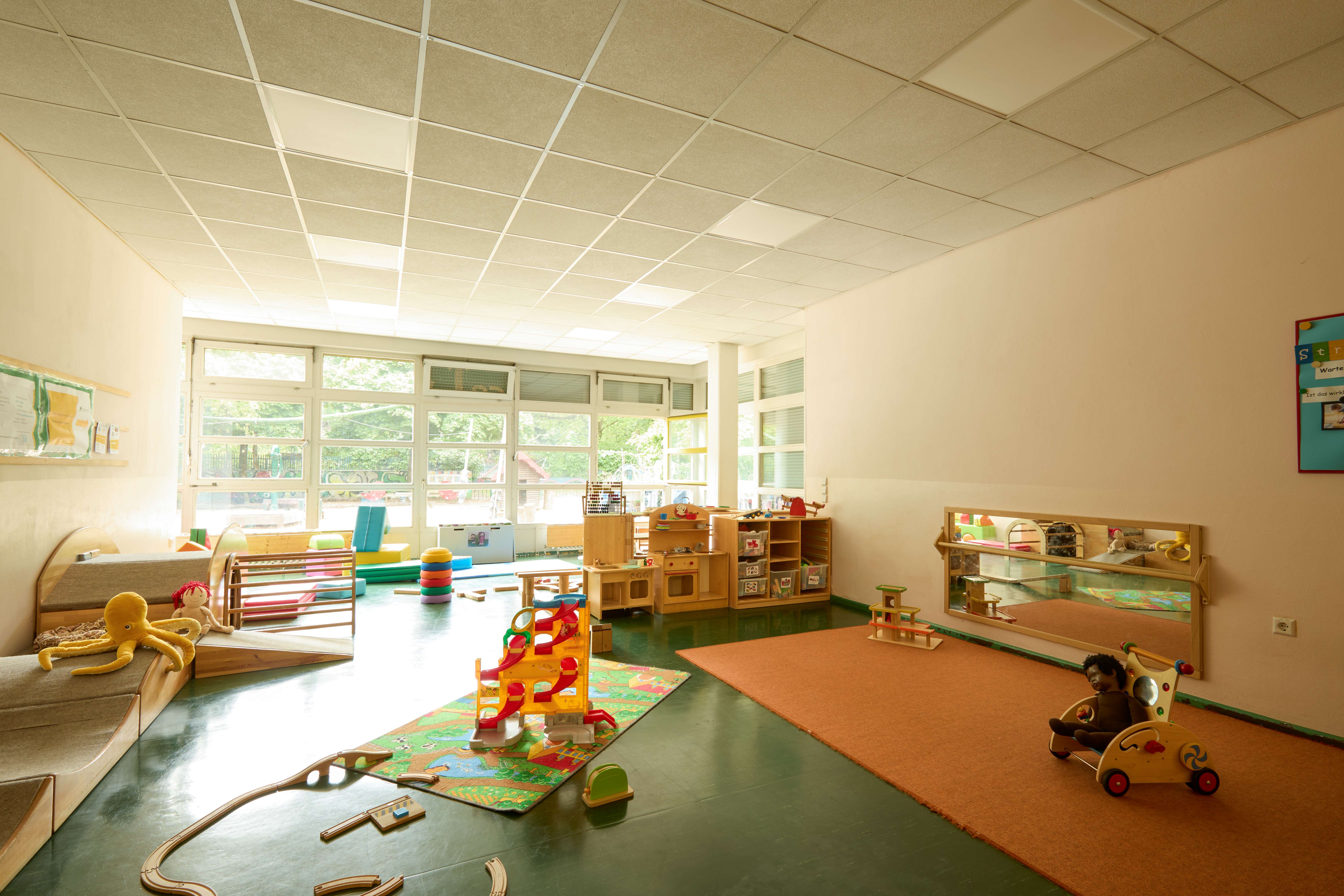 Heller Raum mit großen Fenstern, Spielteppich, Holzspielzeug und Kuscheltieren.