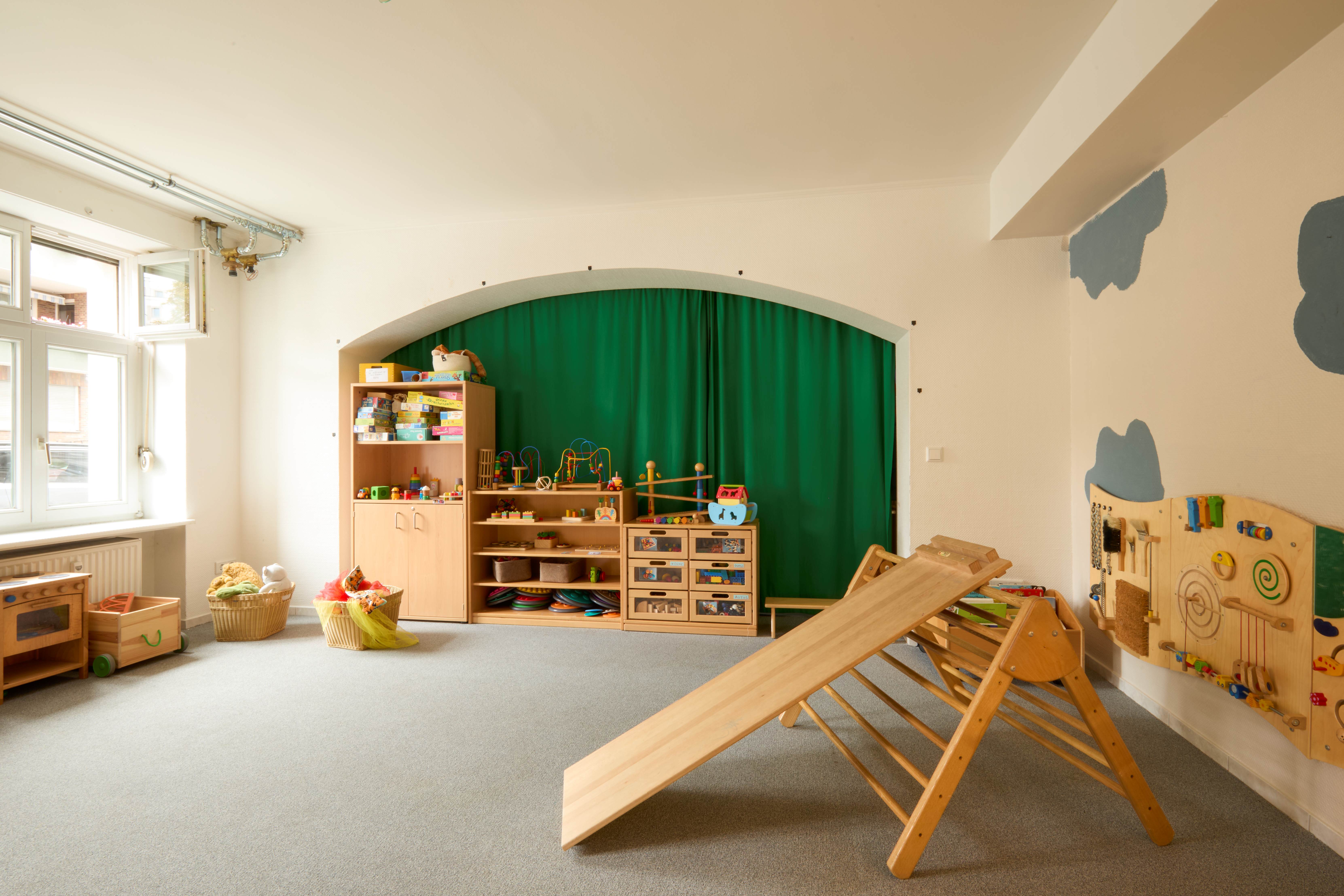 Heller Spielraum für Kinder mit Holzspielzeug: Regale voller Kinderspielzeug vor grünem Vorhang, Kletterdreieck mit Rutsche in der Mitte.