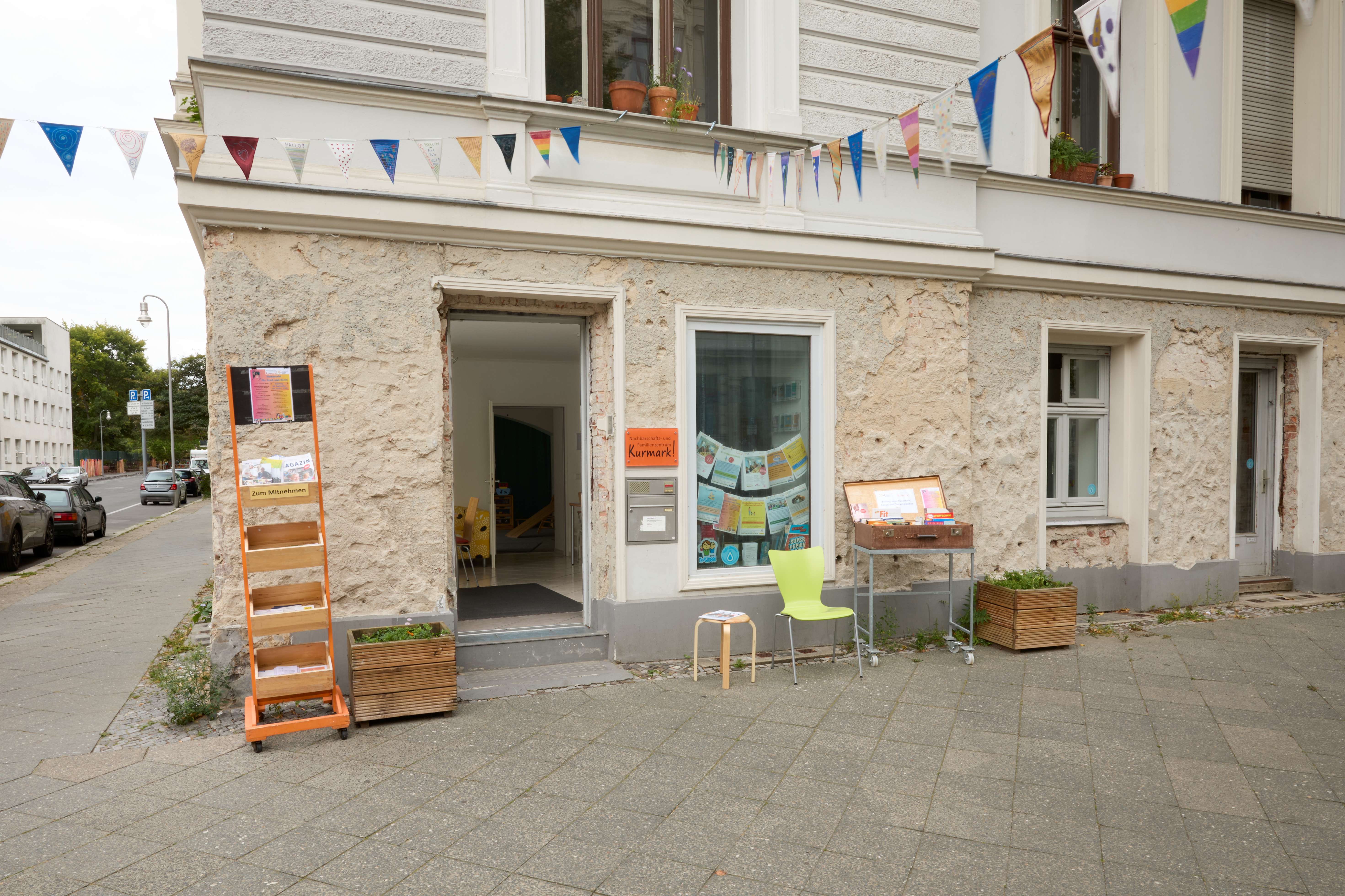 Hausecke mit Eingang zum Nachbarschafts‑/Familienzentrums Kurmark: offene Tür mit kleiner Stufe, Schaufenster mit bunten Flyern, davor grüner Stuhl und Infotisch; darüber Wimpelketten an der Fassade.