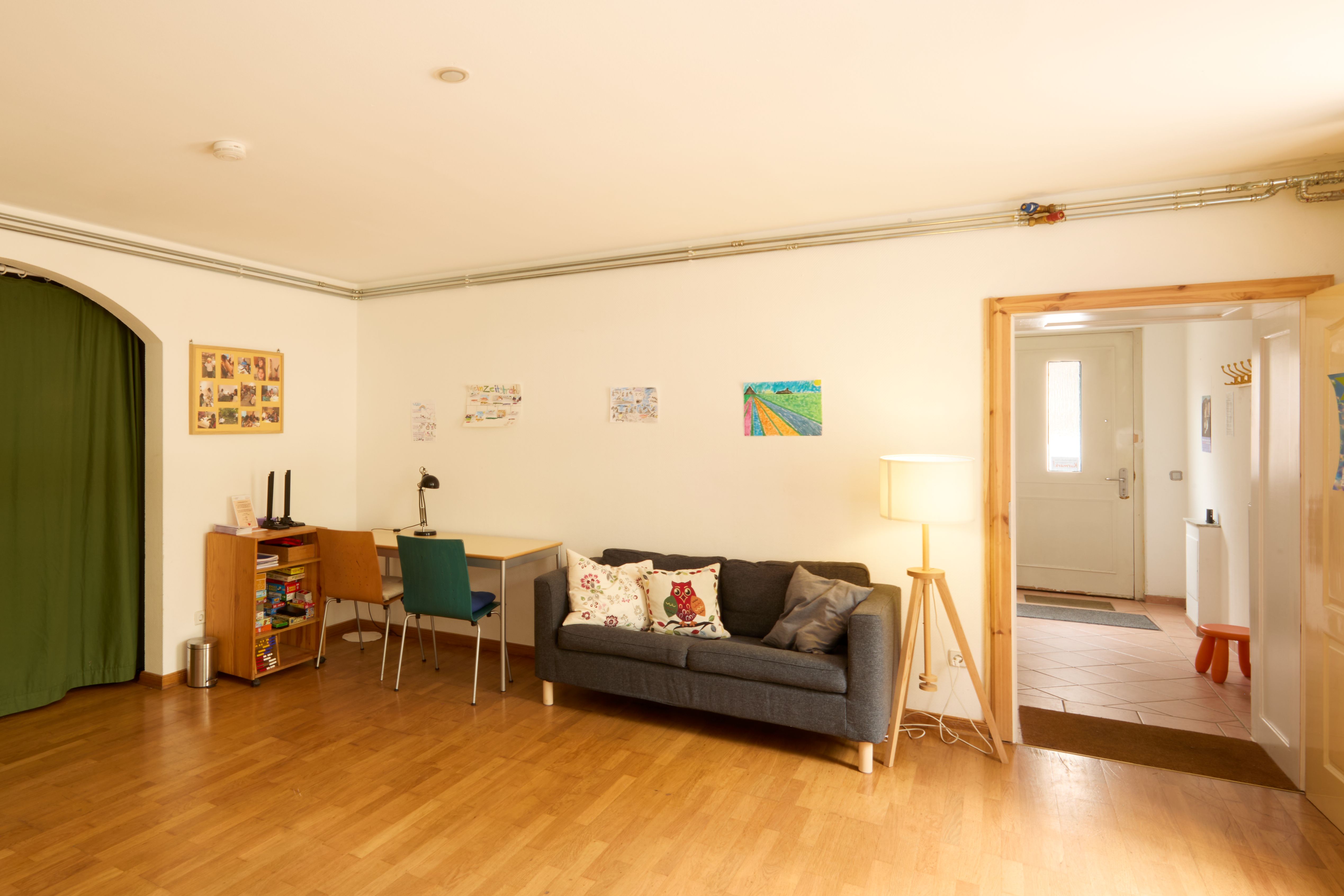 Gemütlicher Raum mit Holzboden, grauem Sofa und Kissen, Stehlampe, kleinem Schreibtisch und Regal; Kinderzeichnungen an der Wand, rechts führt eine Tür in den Flur.
