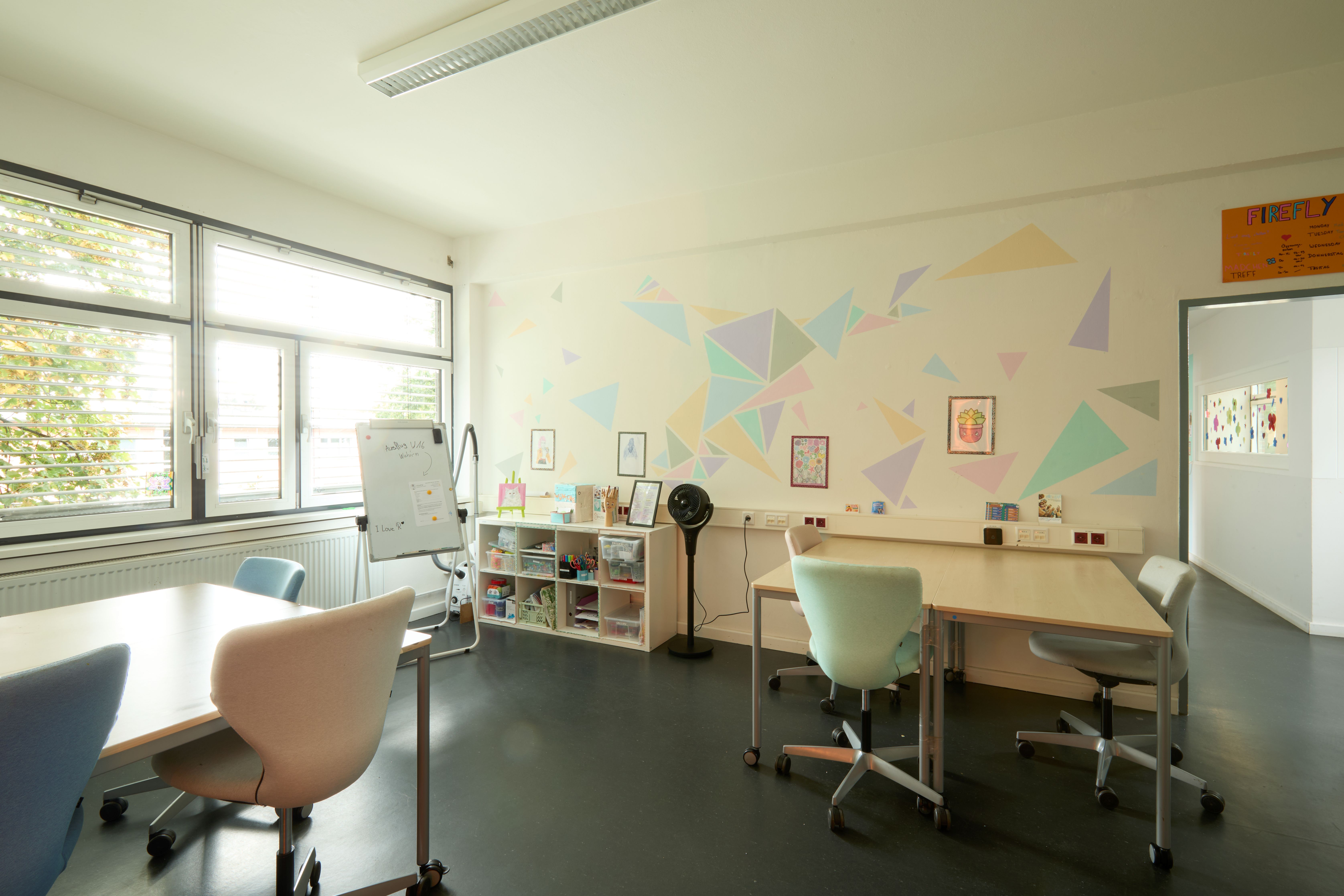 Heller Mehrzweck- und Lernraum mit großen Fenstern, Pastell-Wanddeko in Dreiecken, Tischen auf Rollen und Whiteboard neben Regal mit Bastelmaterial.