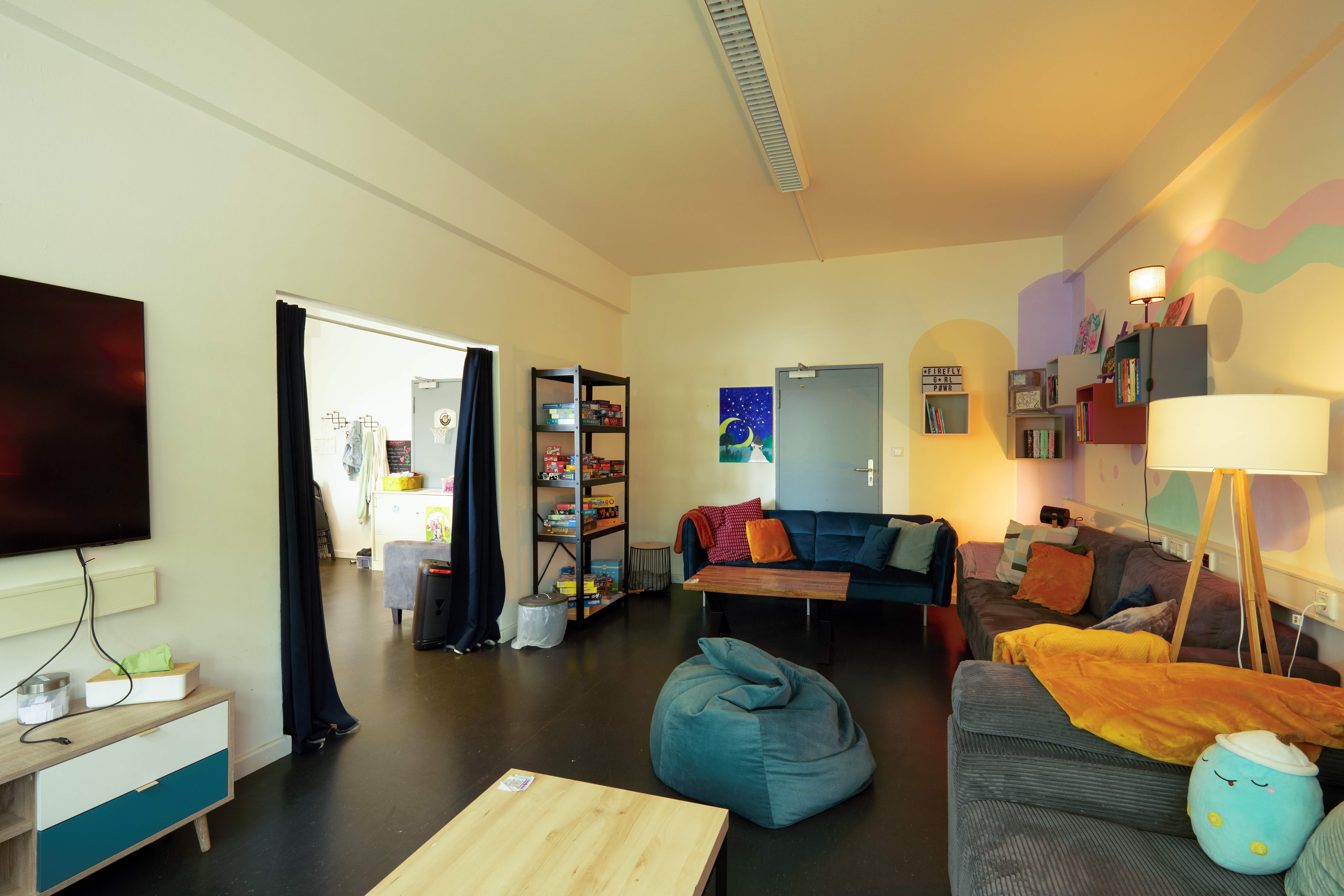 Gemütlicher Jugend- oder Aufenthaltsraum mit Sofas, Sitzsack, Wandregalen mit Spielen, Fernseher und Durchgang in einen weiteren Raum.