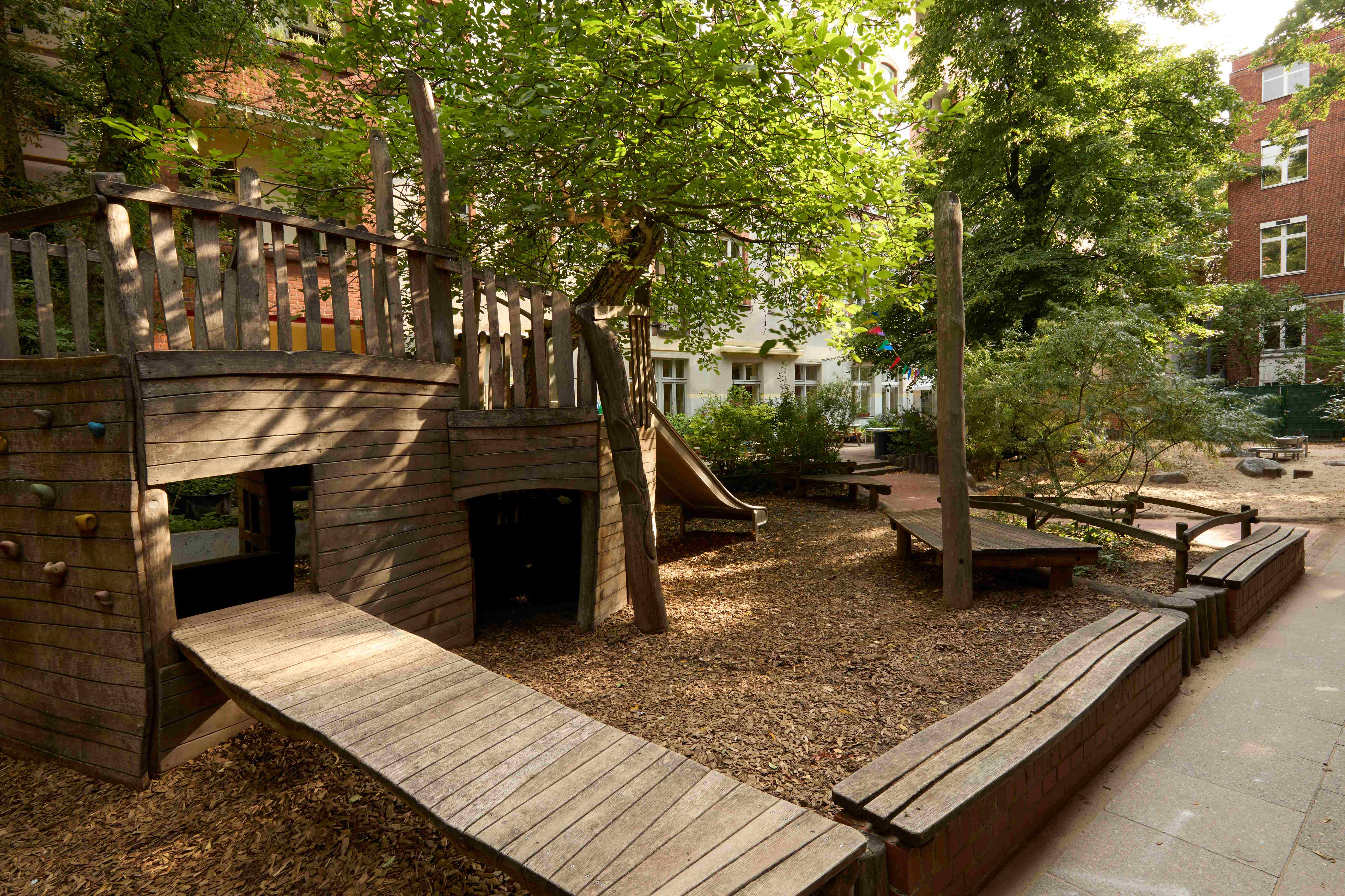 Schattiger Kita‑Hof mit großen Spielplatz aus Holz, Sitzbänken, Bäumen und Sträuchern.