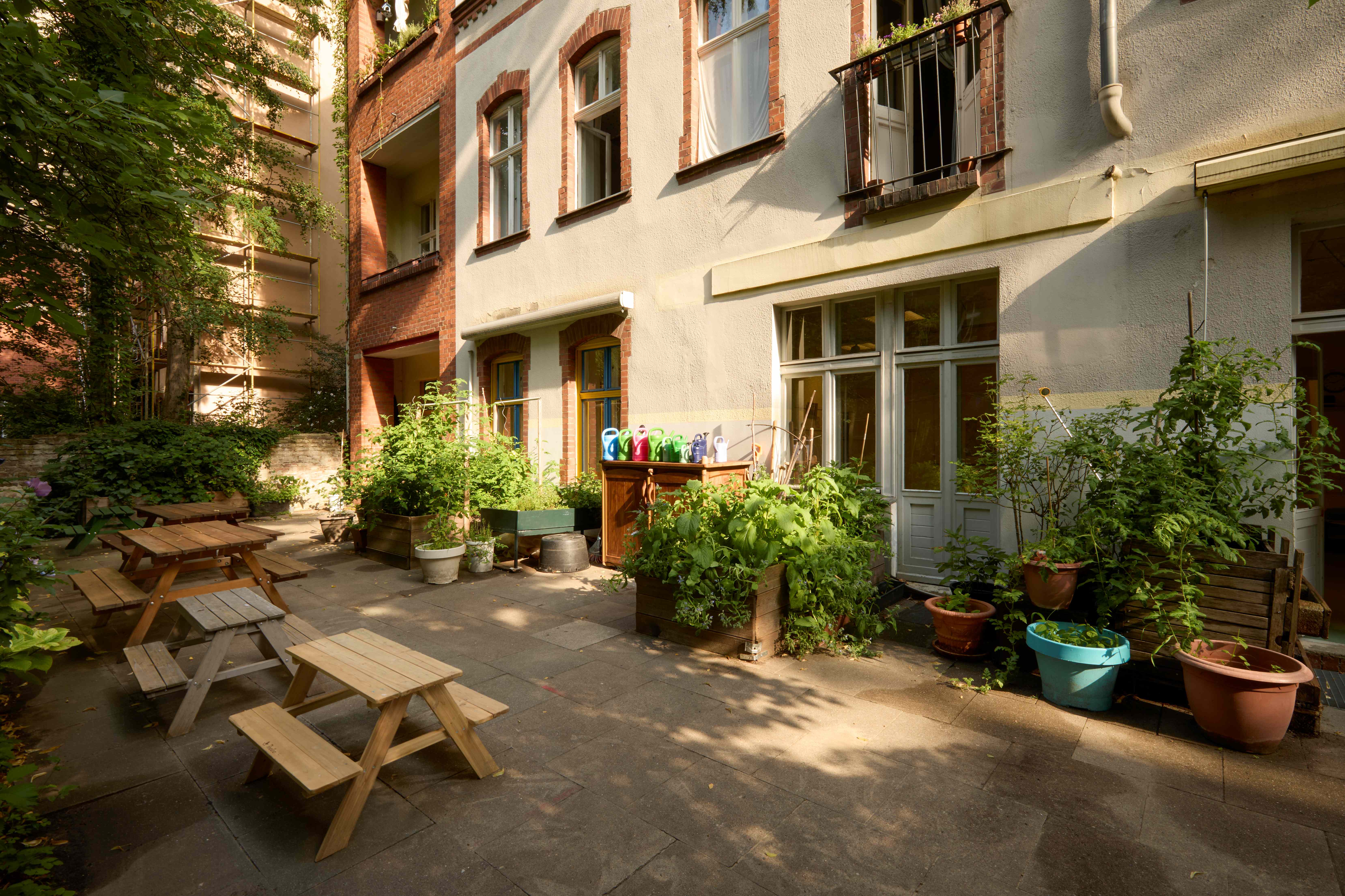 Bepflanzter Innenhof einer Kita mit gepflasterter Terrasse: mehrere kleine Holzbänke und Picknicktische, entlang der Hauswand viele Hochbeete und Pflanzkübel mit Gemüse und Kräutern; Gießkannen auf einem Schrank, dahinter Altbaufassade mit Fenstern und Türen.