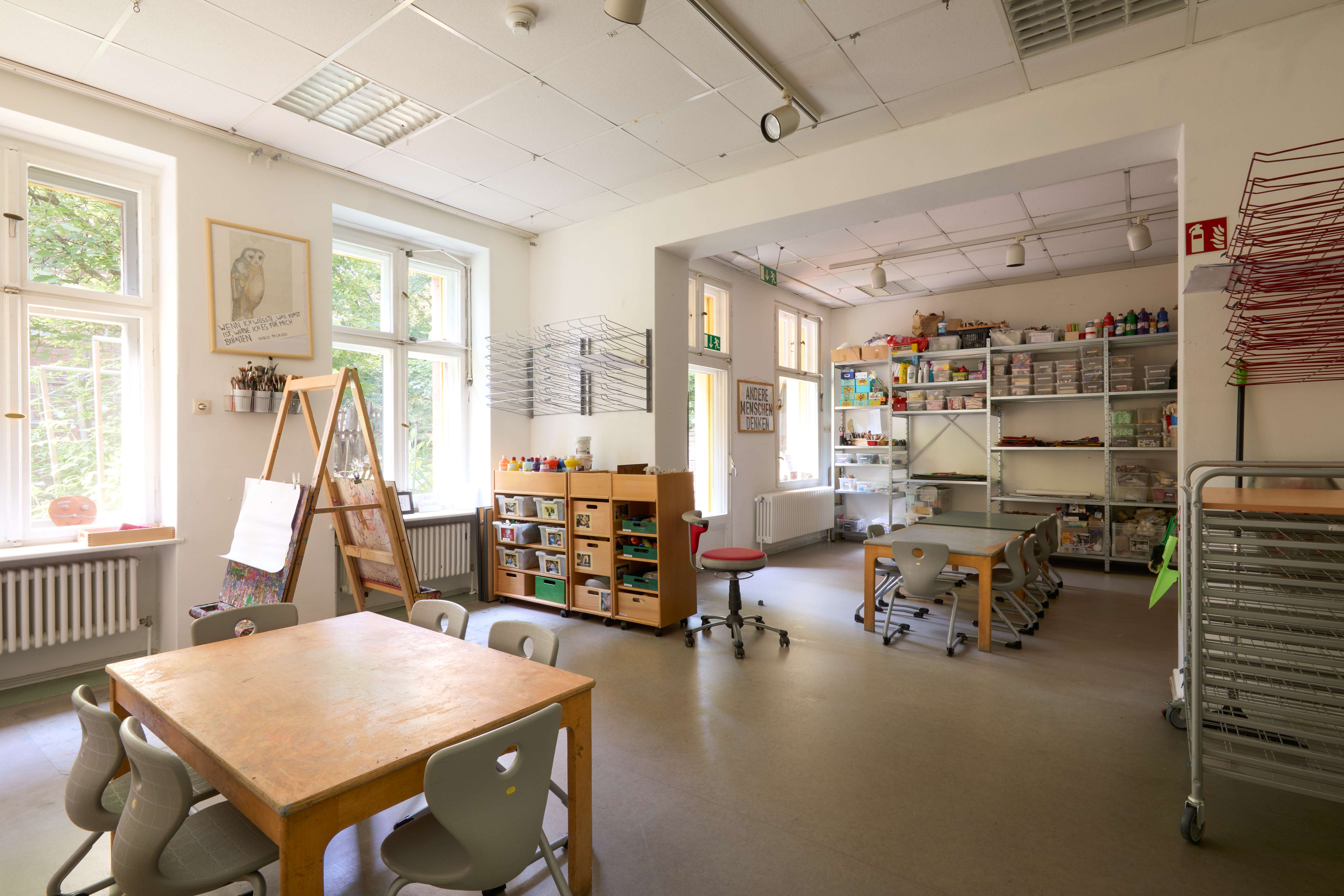 Großes Atelier der Kita mit Arbeitstischen und Staffeleien, links helle Fenster; im hinteren Raum hohe Regale mit sortierten Kunstmaterialien.