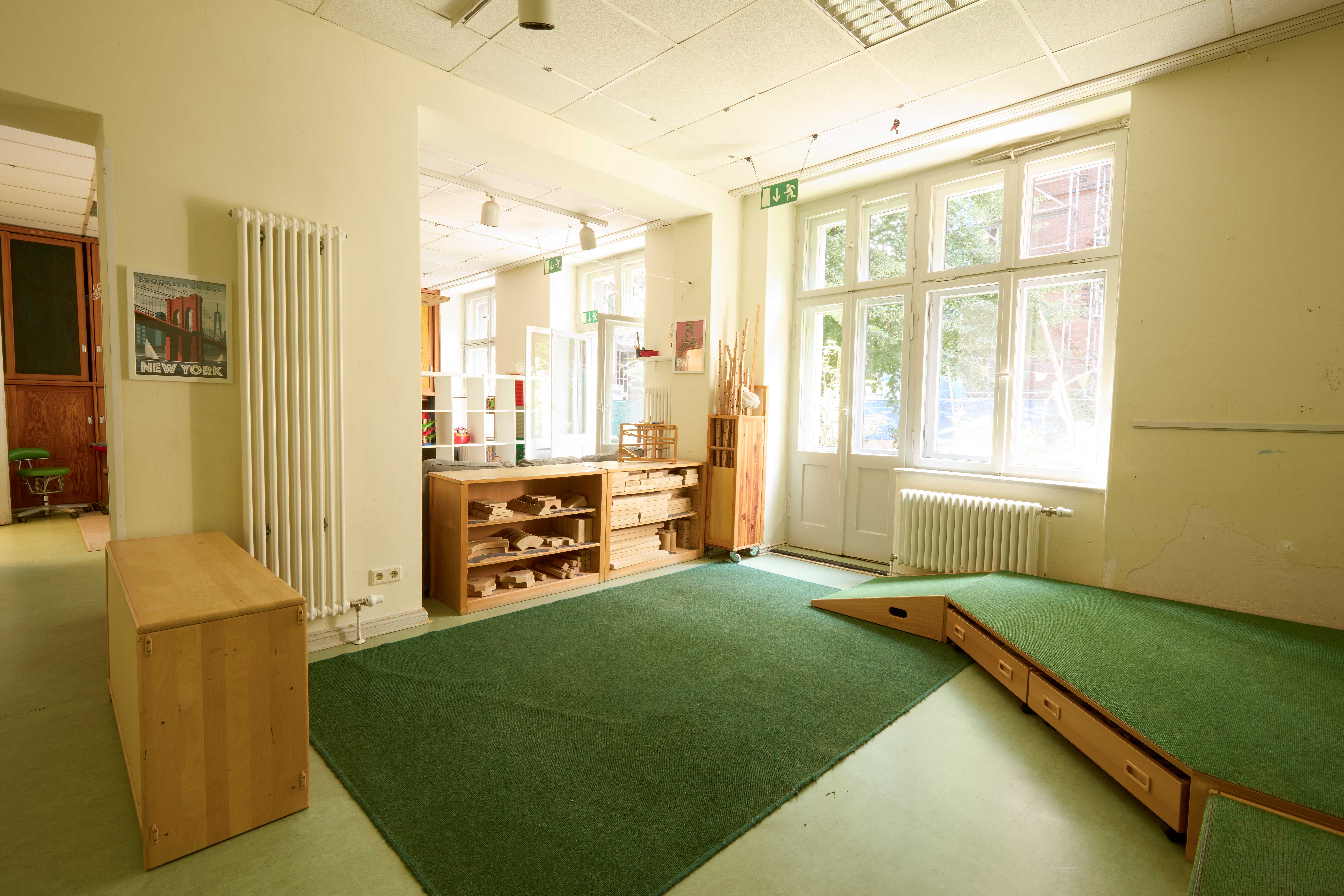 Heller Kita‑Bauraum mit grünem Teppichpodest und Schubladen, großen Fenstern und Tür zum Hof; an der Wand offene Holzregale mit Baumaterial, daneben Materialwagen.