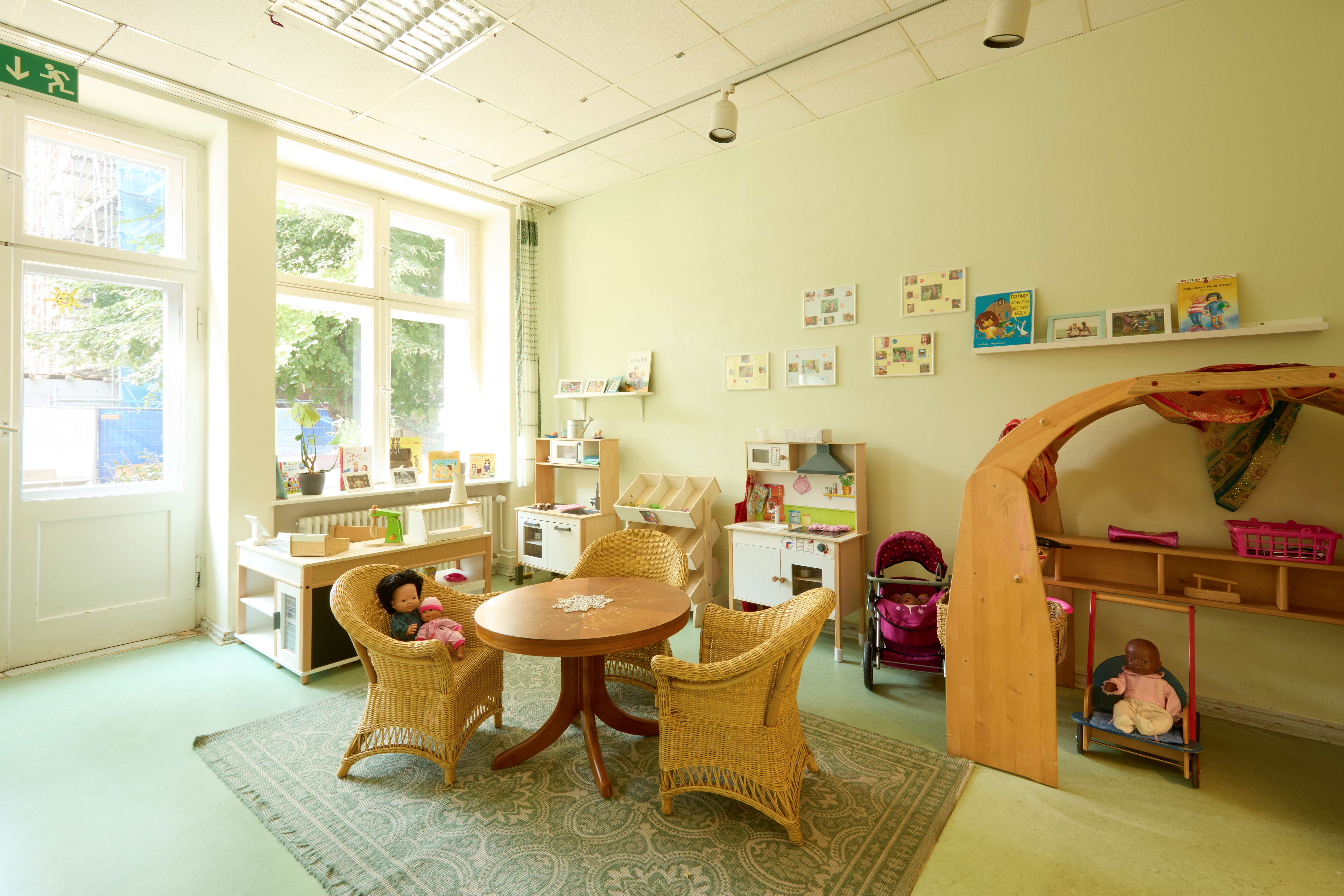 Freundlicher Rollenspielraum der Kita: große Fenster, runder Holztisch mit drei Korbstühlen auf Teppich, Spielküchen und Regale an der Wand, Puppenwagen und Holzspielbogen mit Tüchern; insgesamt warmes, wohnliches Ambiente.