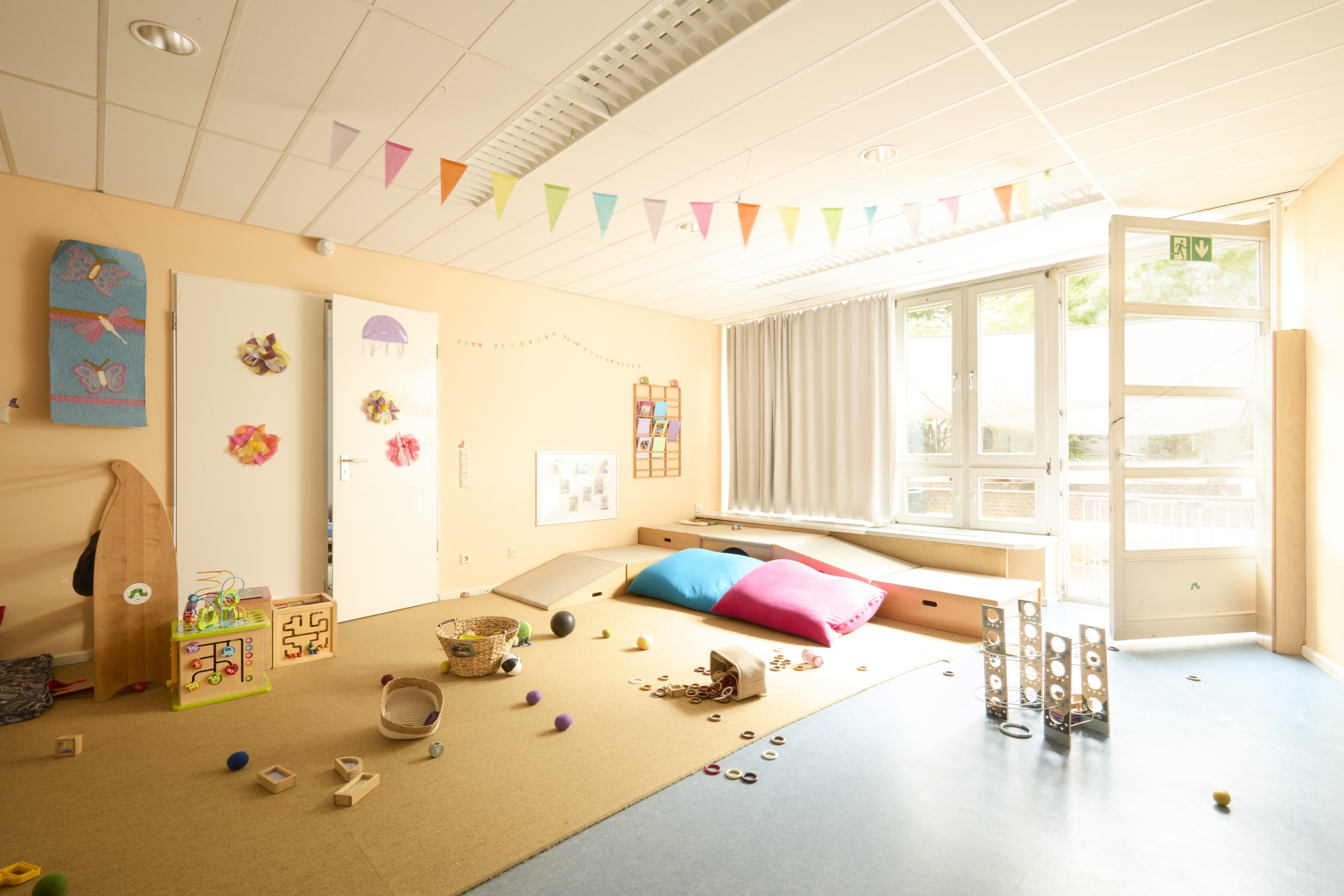 Heller Gruppenraum mit großen Fenstern, Tischen und kleinen Stühlen. An der Decke hängen bunte Girlanden; auf dem Boden liegen Spielsachen.