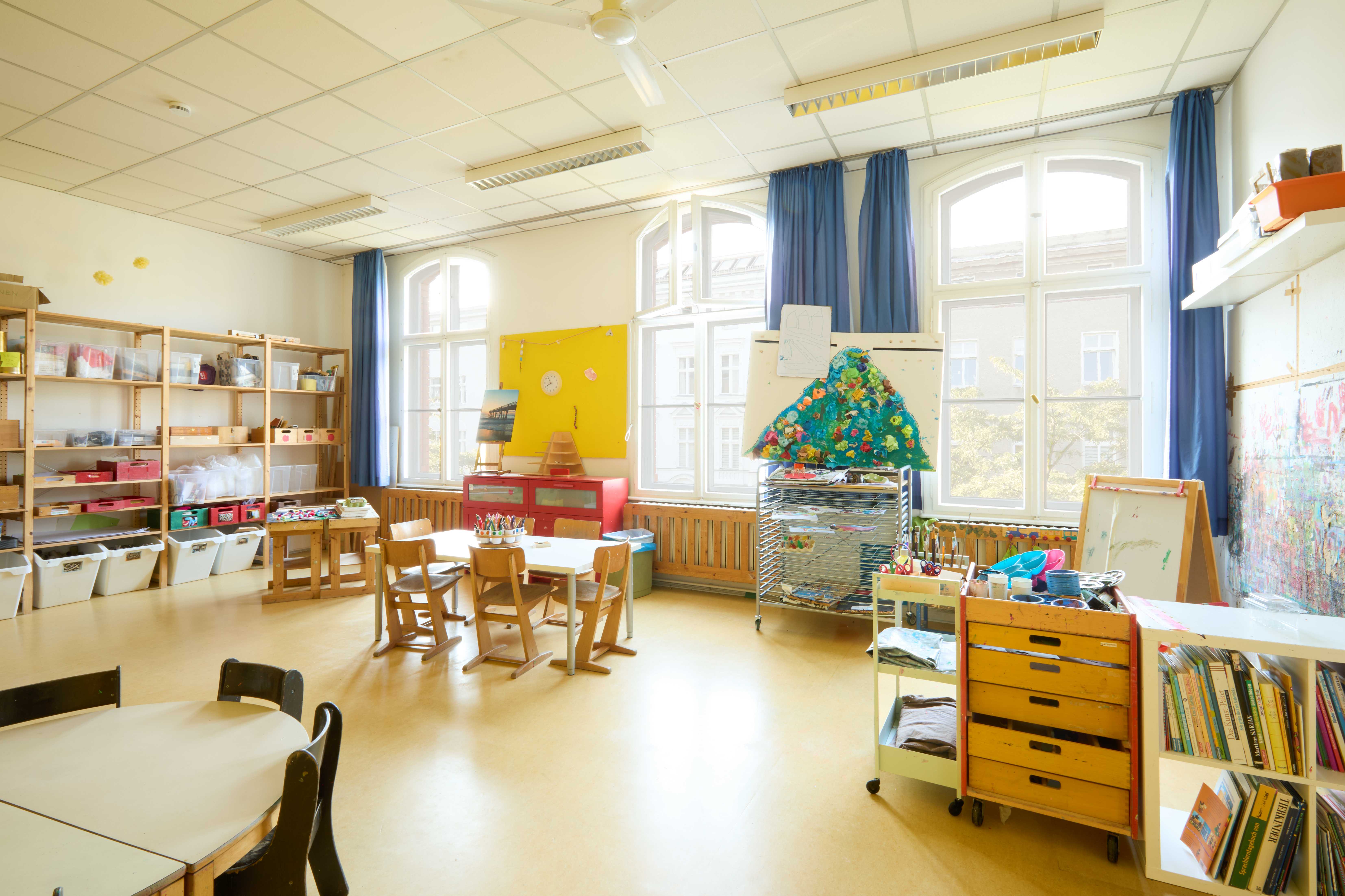 Großes Atelier mit hohen Fenstern und viel Material: Regale mit Kisten, Maltische, Staffelei; gelbe Pinnwand und mobile Wagen mit Kreativwerkzeugen.