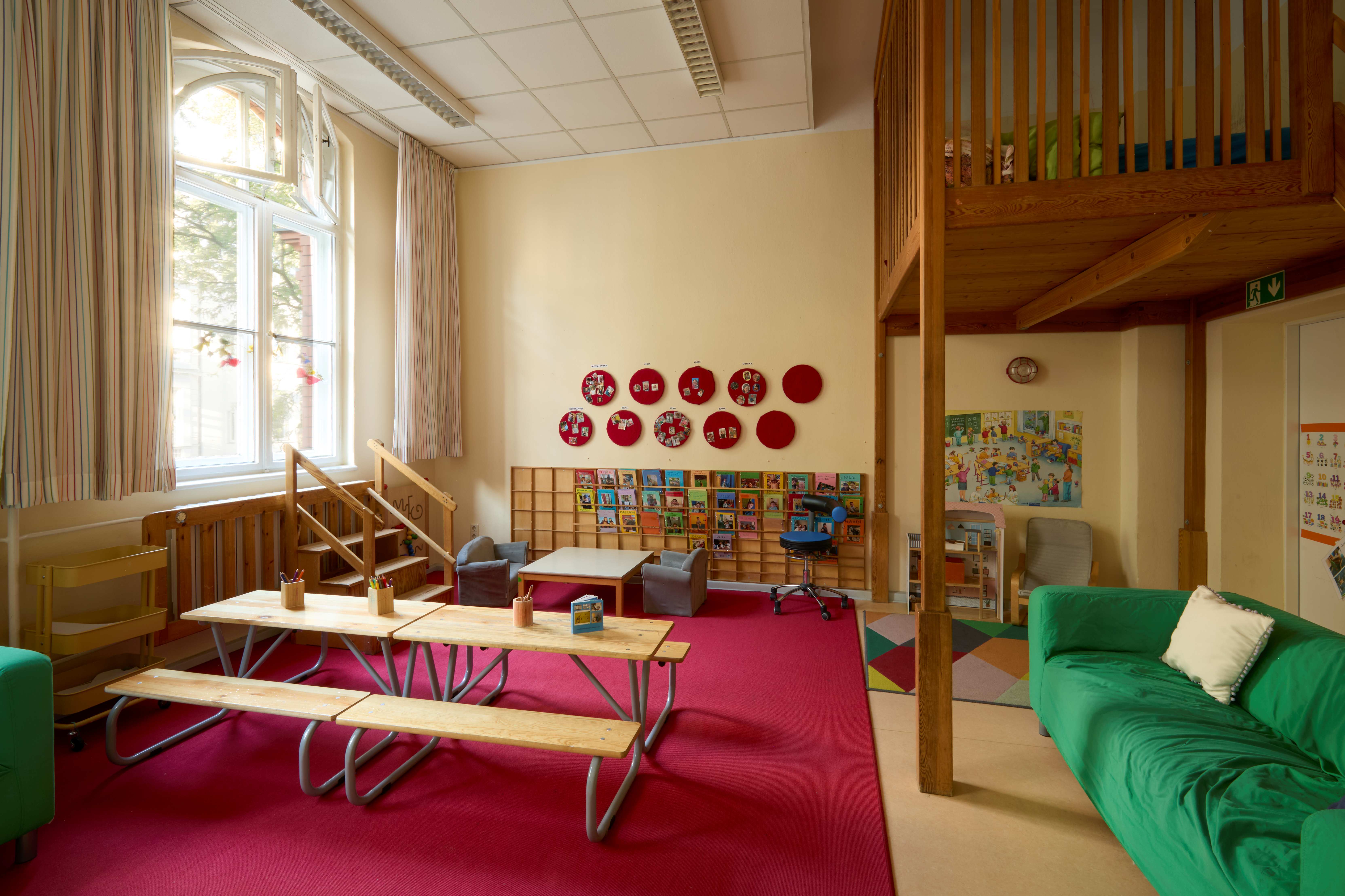 Gemütlicher Begrüßungsraum mit rotem Teppich, niedrigen Tischen und Sitzgruppen; Bilder‑ und Bücherwand, große Fenster und eine erhöhte Ebene aus Holz neben einem grünen Sofa.