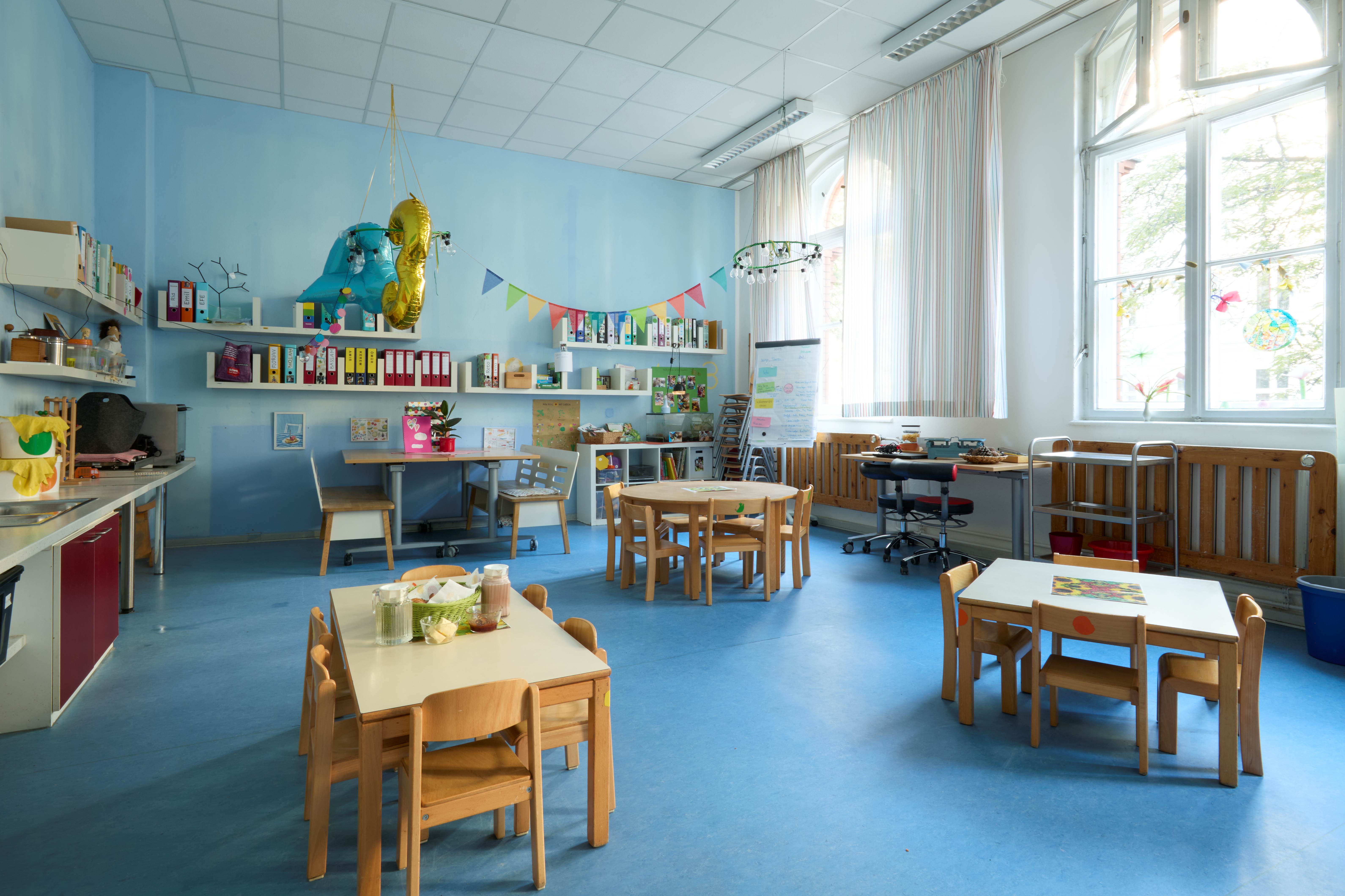 Freundlicher Essbereich mit hellblauen Wänden, Kinder‑Tischen und -Stühlen; Küchenzeile links, Regalen, Deko‑Girlanden, große Fenster mit Tageslicht.
