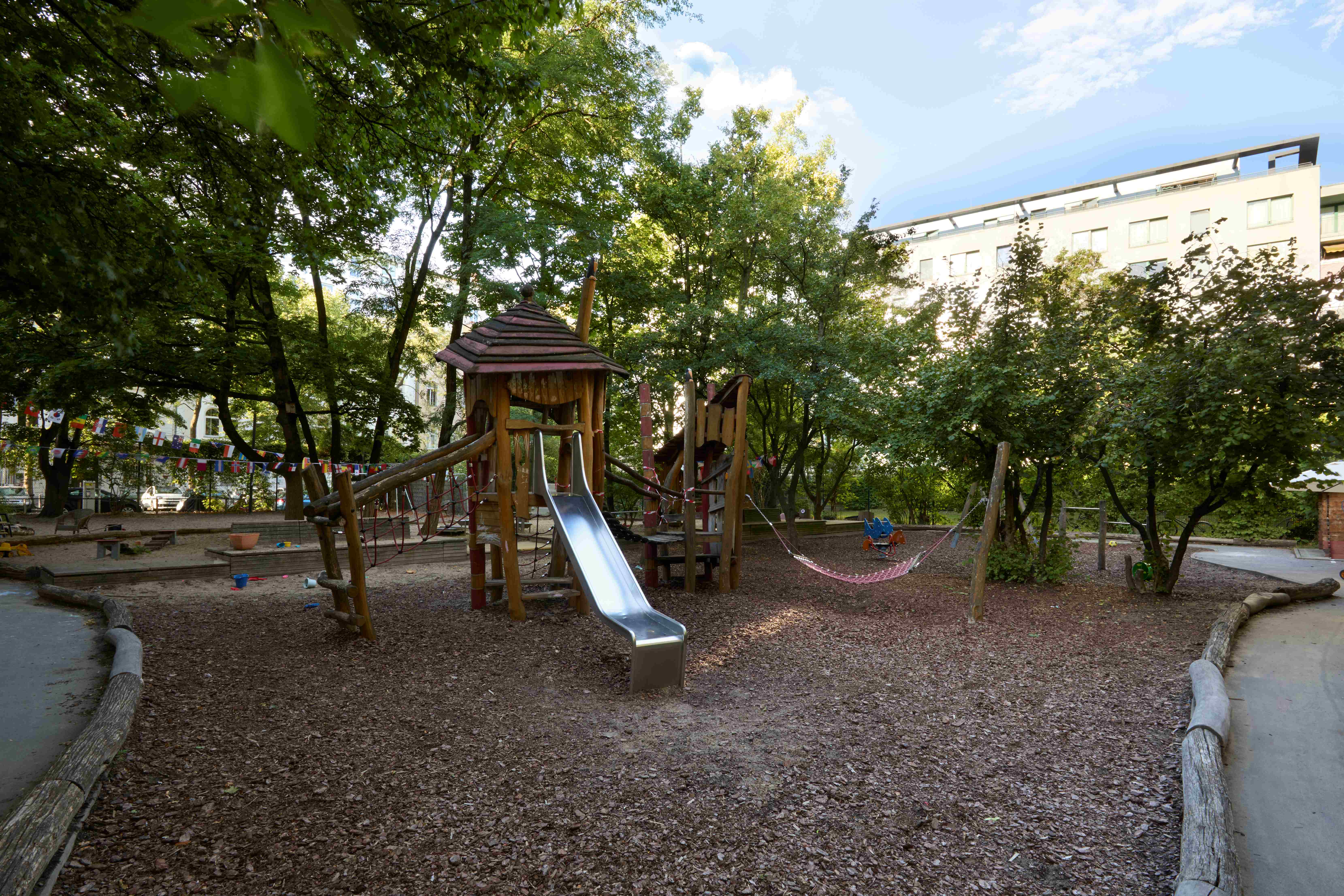 Schattiger Kita‑Spielplatz mit Holz‑Kletterturm, Seilbrücken und Rrutsche umgeben von vielen Bäumen und bunten Wimpelketten.