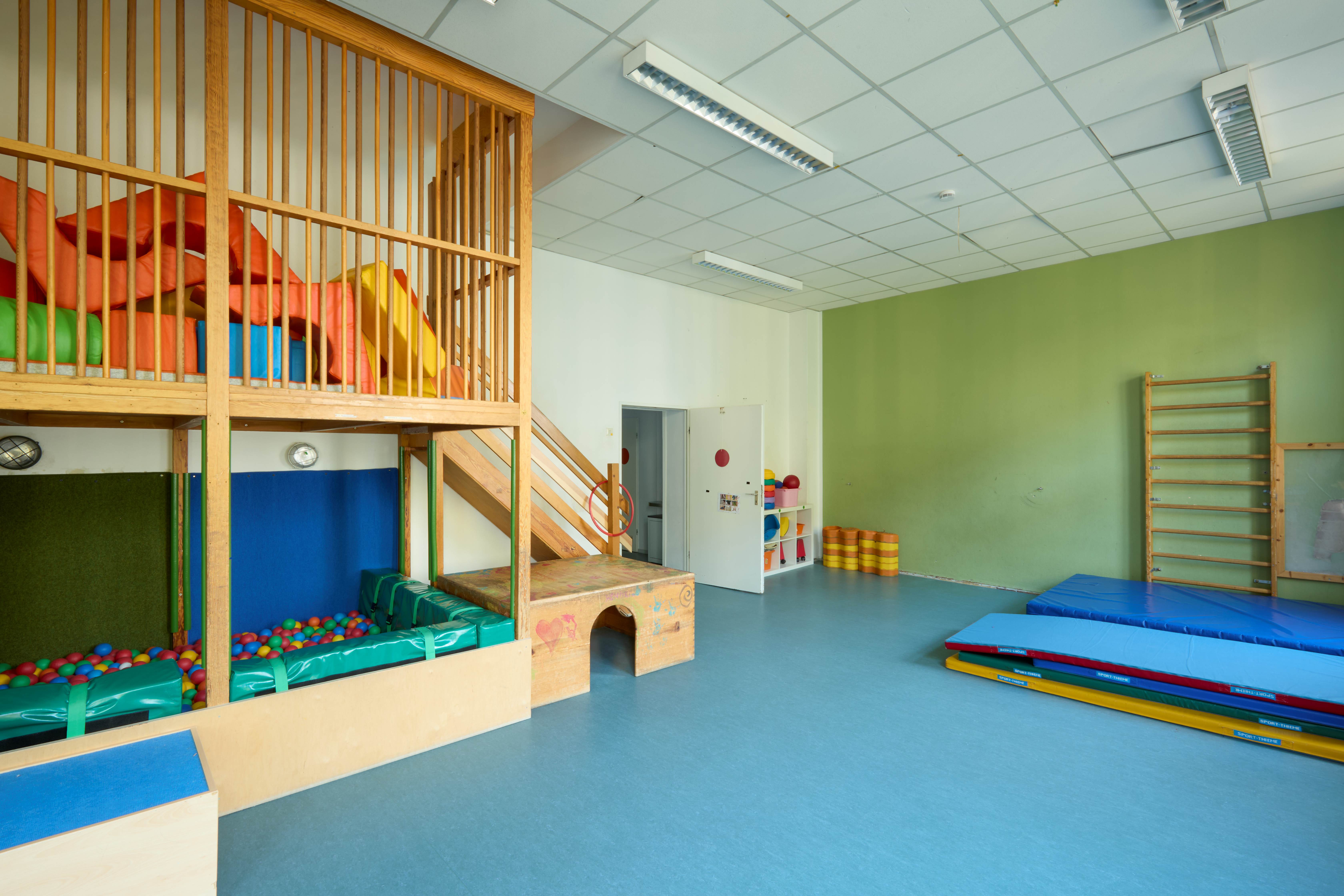 Heller Bewegungsraum mit Kletterpodest, Treppe und zwei Bällebädern unten; blaue Turnmatten, Sprossenwand und Regale mit bunten Sportmaterialien.
