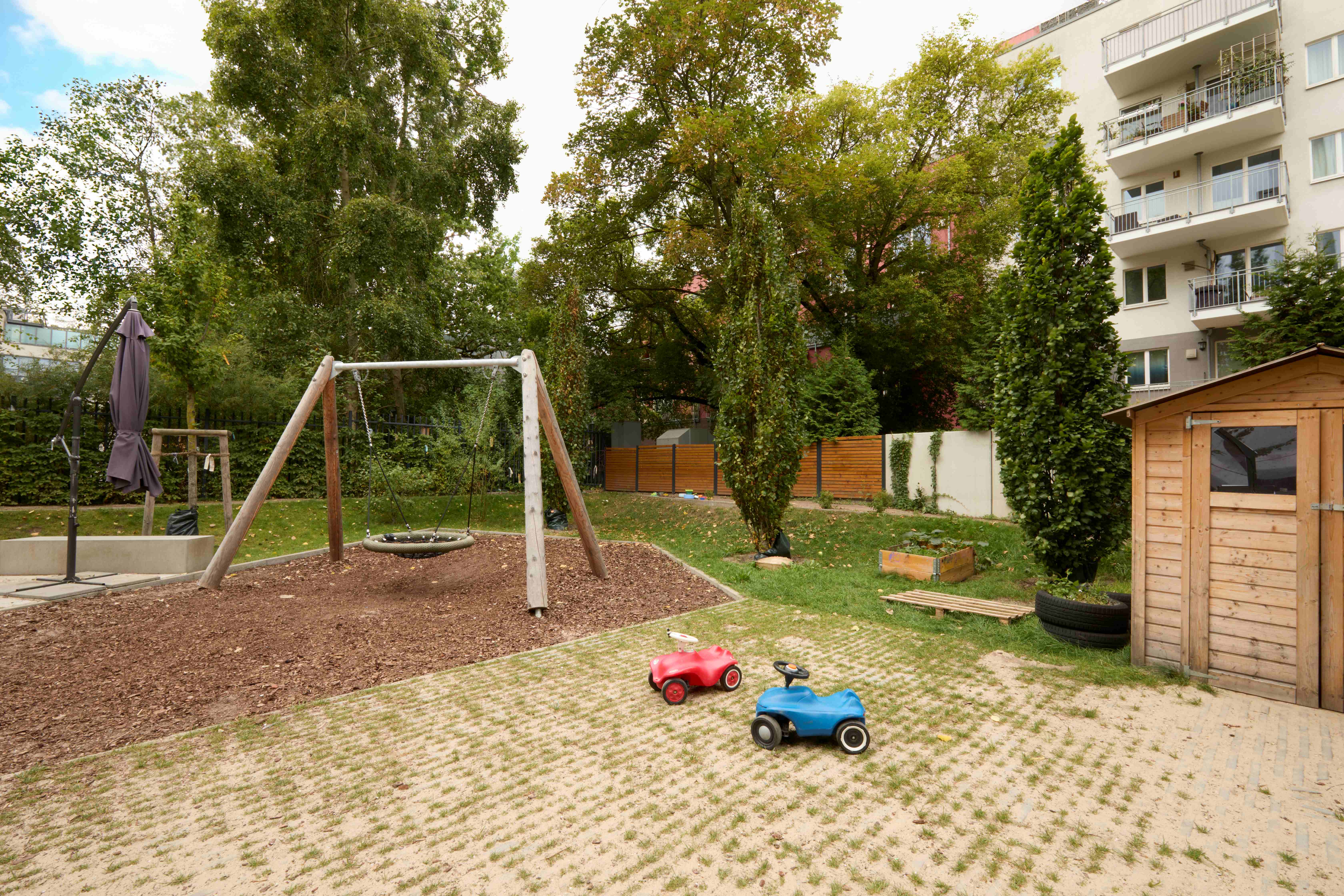 Grüner Kita‑Garten mit Schaukel, zwei Rutschautos, einem Sonnenschirm, einem Hochbeet und einem kleinen Holzschuppen; umgeben von Bäumen.