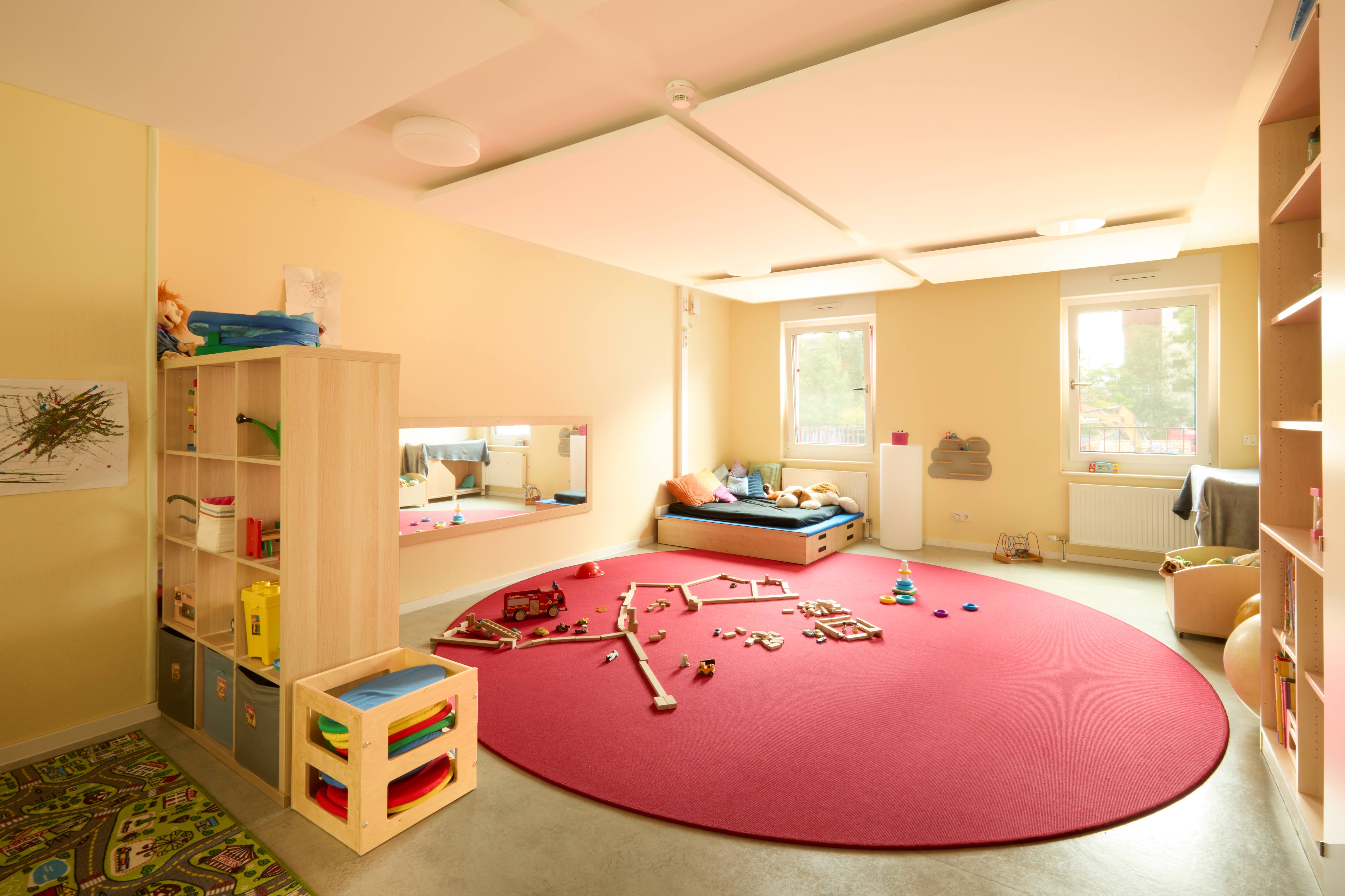 Gemütlicher Gruppenraum mit großem rotem Rundteppich, Spielmaterial auf dem Boden und Kuschelecke mit Matratze am Fenster; offene Regale und Spiegel in Kinderhöhe.