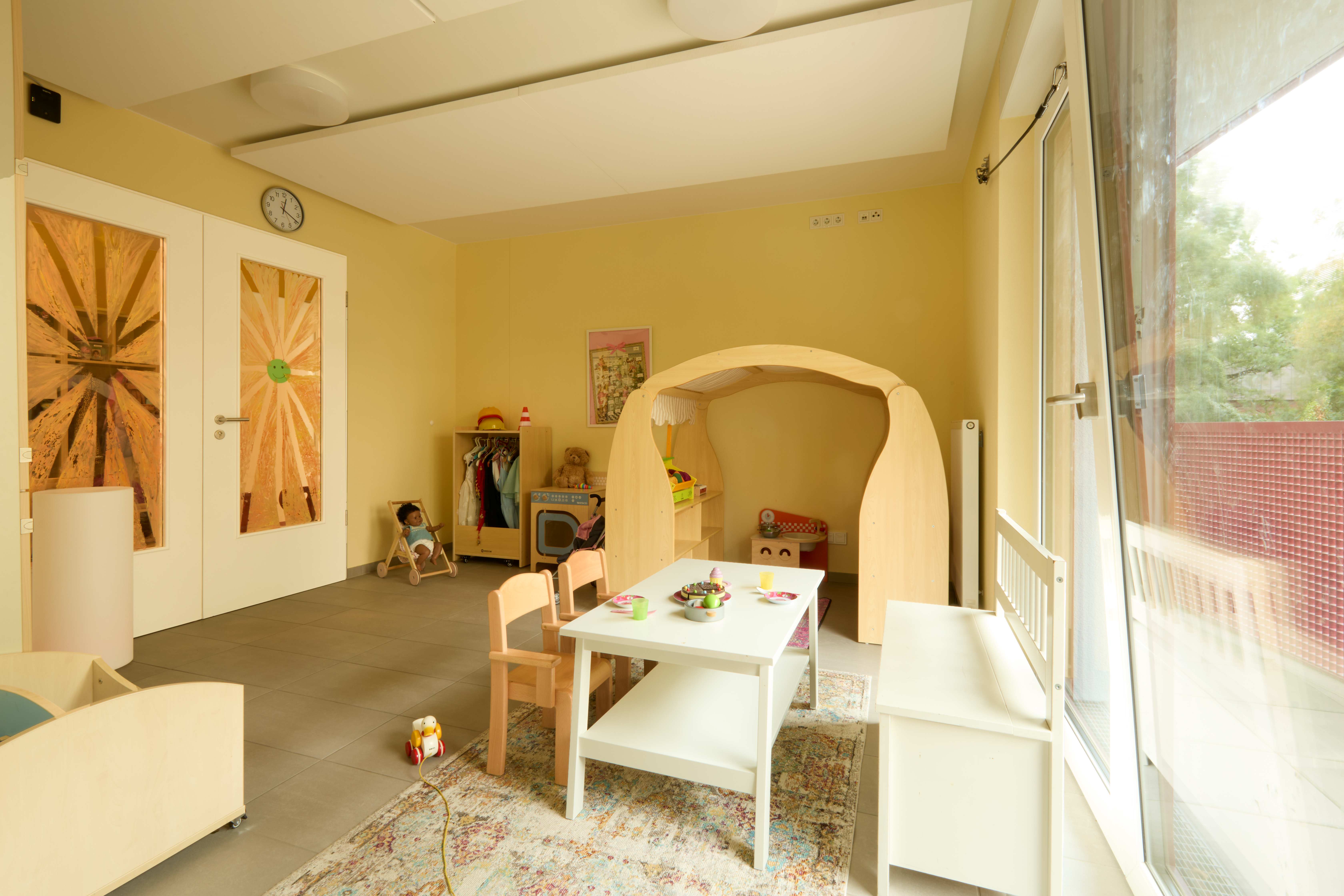  Spielraum mit hellem Holzhäuschen, Puppenküche und Verkleidungsecke; kleiner weißer Tisch mit Stühlen auf Teppich, große Fenster zum Balkon.