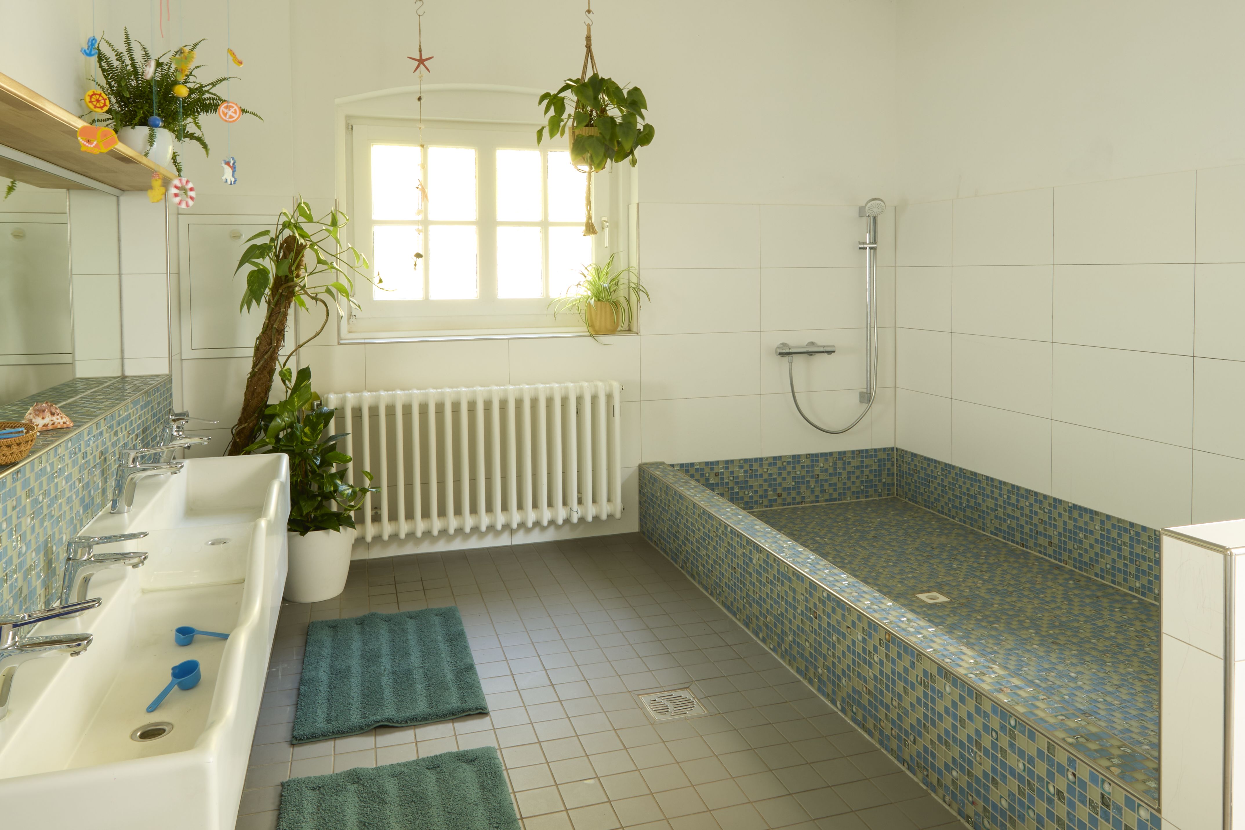 Ein heller Waschraum ist mit weißen Waschbecken und Fliesen ausgestattet. Rechts befindet sich eine geflieste Dusch- oder Badebereich, und auf dem Boden liegen türkisfarbene Matten.