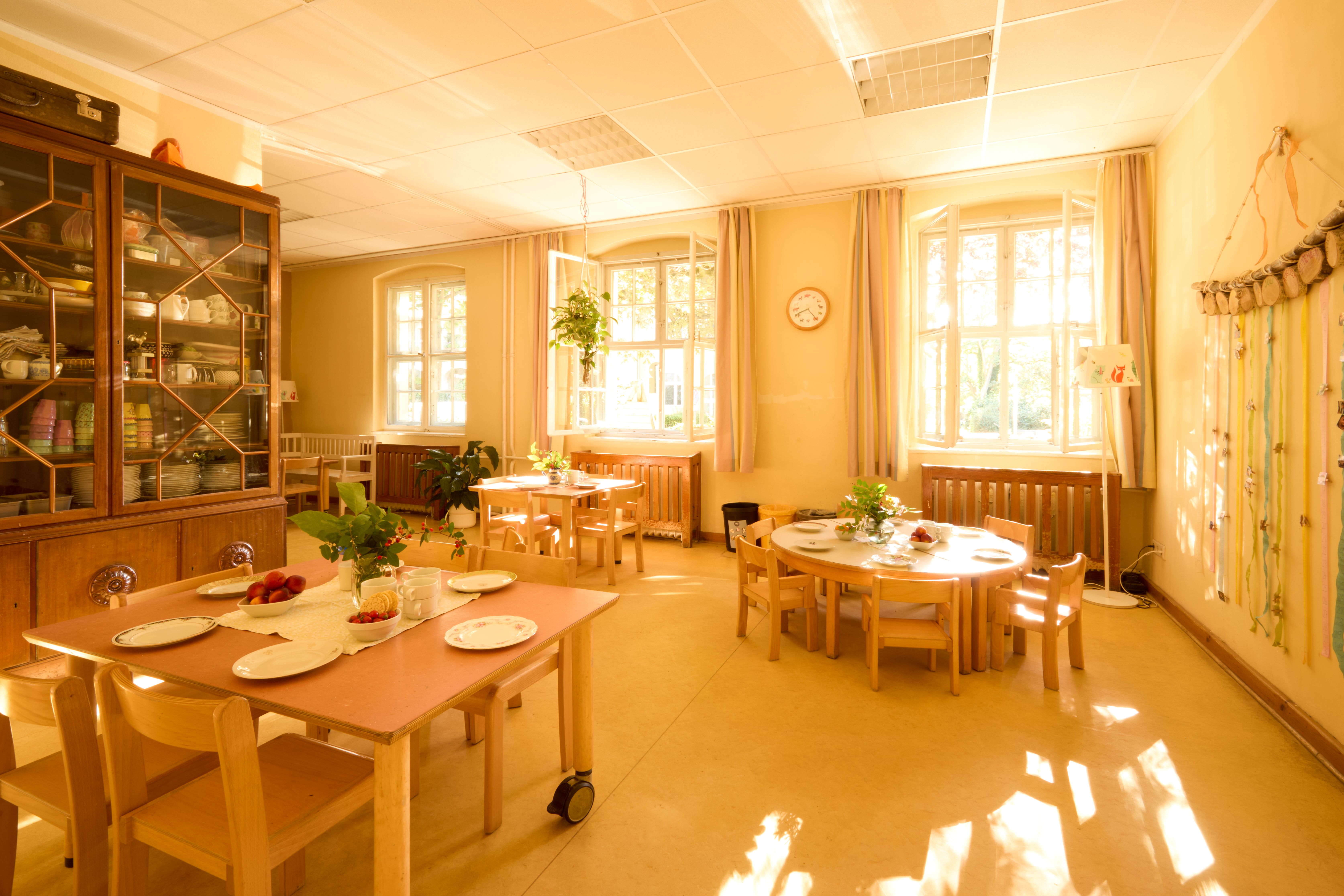 Ein heller Essbereich ist mit langen Holztischen und Stühlen eingerichtet; auf den Tischen steht Geschirr und große Fenster lassen Tageslicht herein.