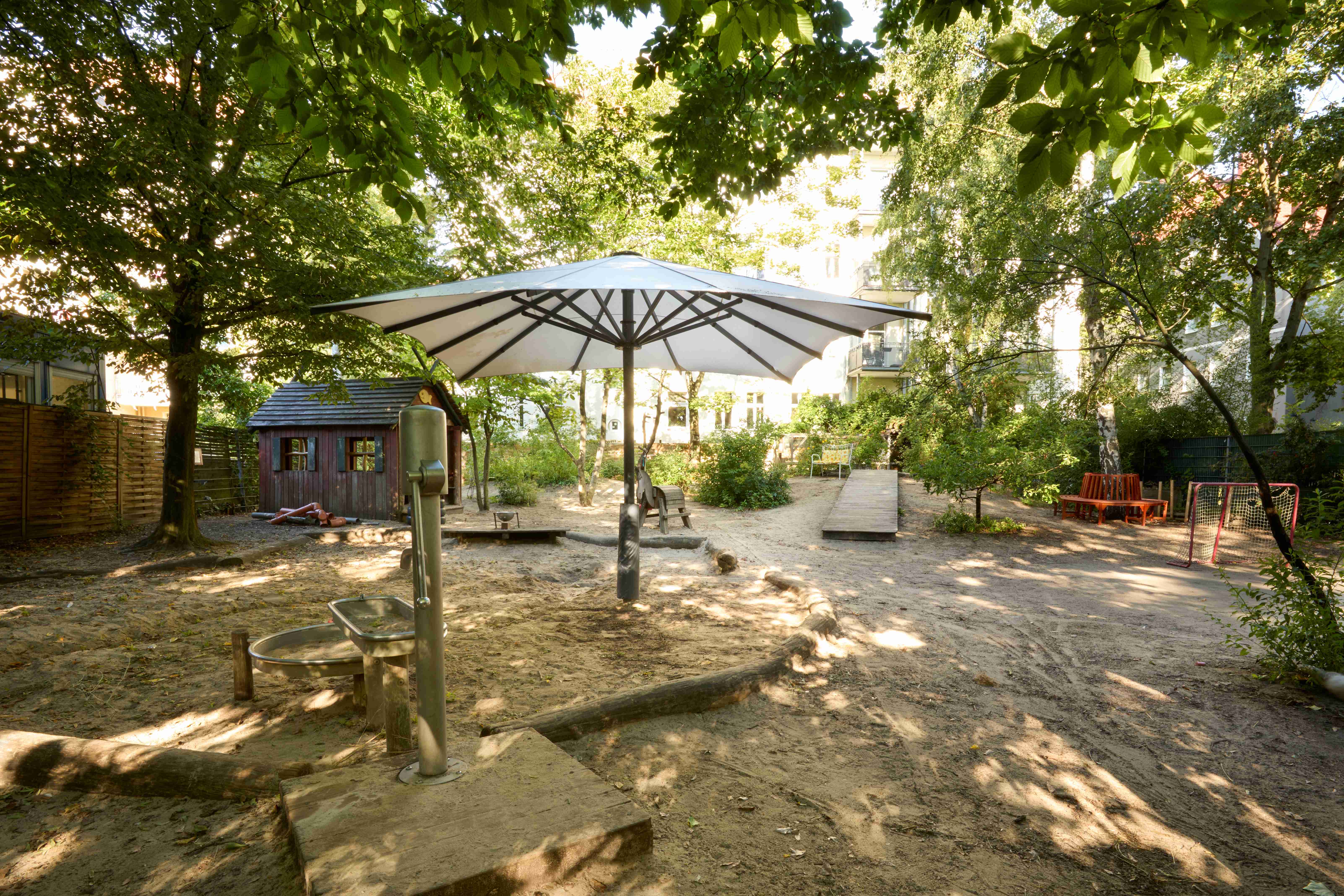 Das dichte grüne Blätterdach eines Baumes spendet Schatten über einem Sitzplatz mit Wasserspiel, einem Tor, Sitzbänken und einer kleinen Holzhütte.