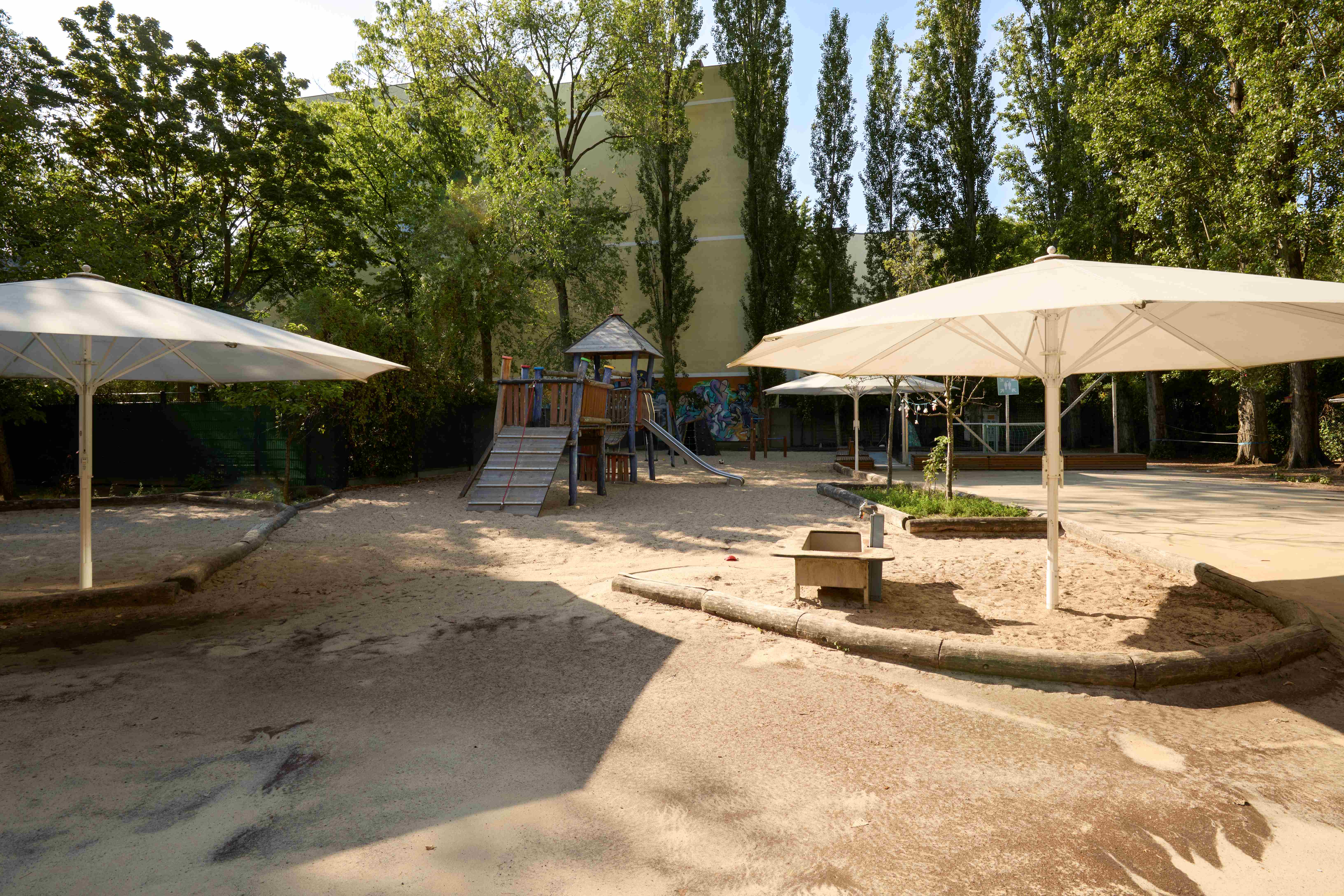 Großer Kita‑Außenspielplatz mit weitläufigen Sandflächen und mehreren weißen Sonnenschirmen; mittig ein hölzernes Klettergerüst mit Rutschen und Kletterrampen, umgeben von Bäumen. Rechts kleine Beete und offene Spielflächen, links schattige Bereiche.