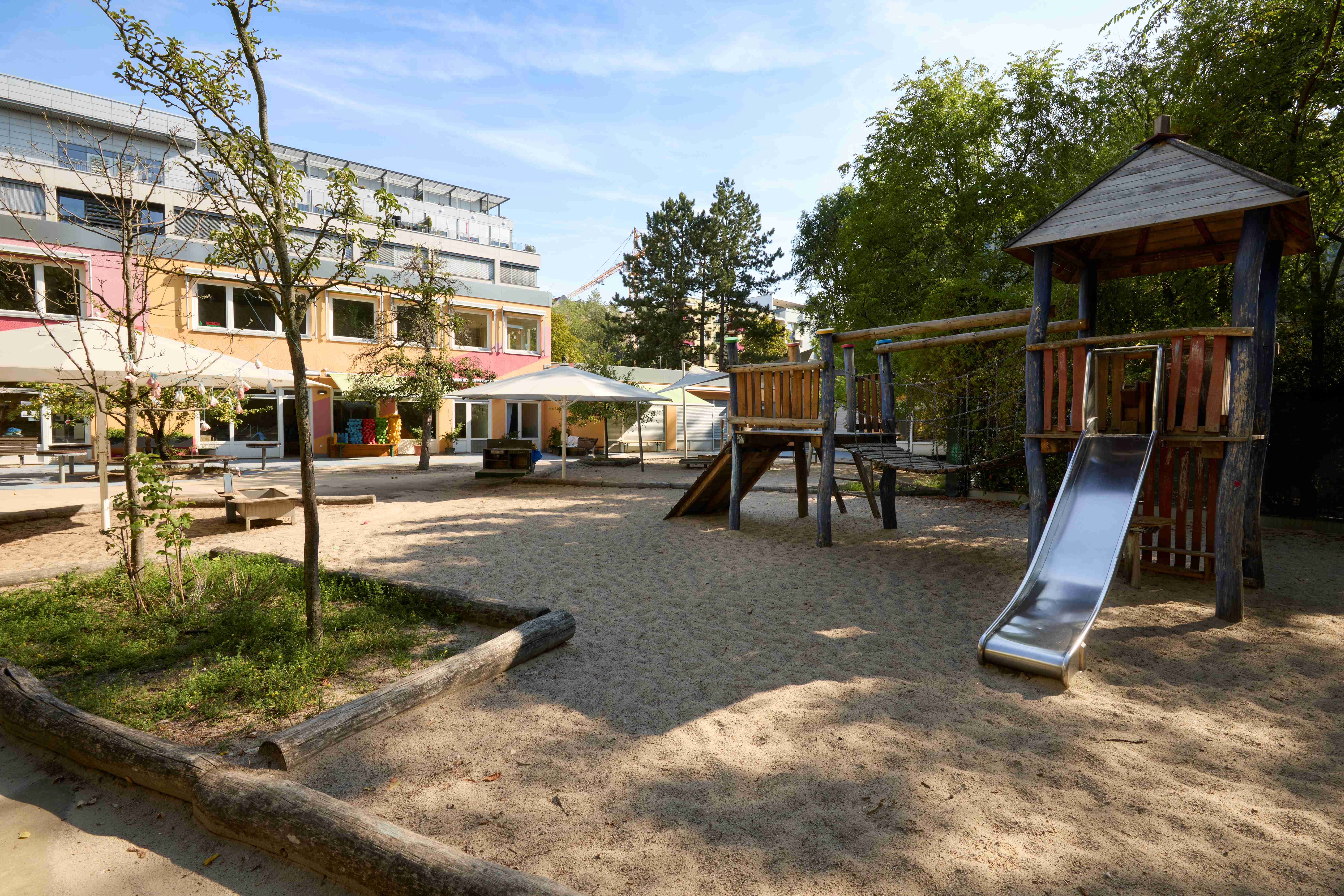 Weitläufiger Kita‑Garten mit Sandflächen: rechts ein hölzernes Klettergerüst mit Netzbrücke und Rutsche, links Beete und Bäume; im Hintergrund das farbige Kita‑Gebäude mit großen Fenstern und Sonnenschirmen.