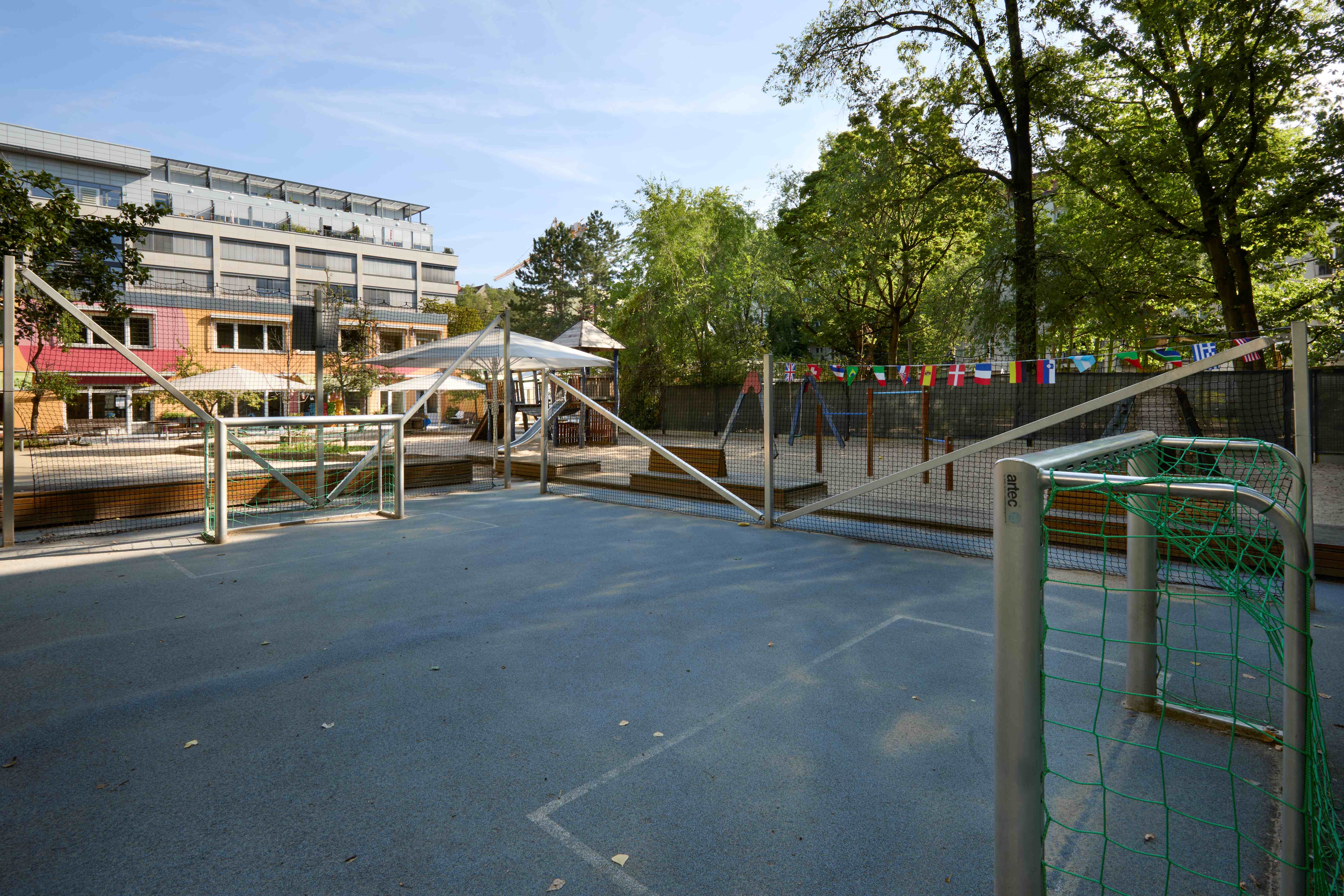 Kleines eingezäuntes Sportfeld und kleinem Tor im Vordergrund; dahinter Zaun mit Wimpelgirlande, rechts Schaukeln im Sandbereich; links Kita‑Gebäude mit Sonnenschirmen und Bäumen drumherum.