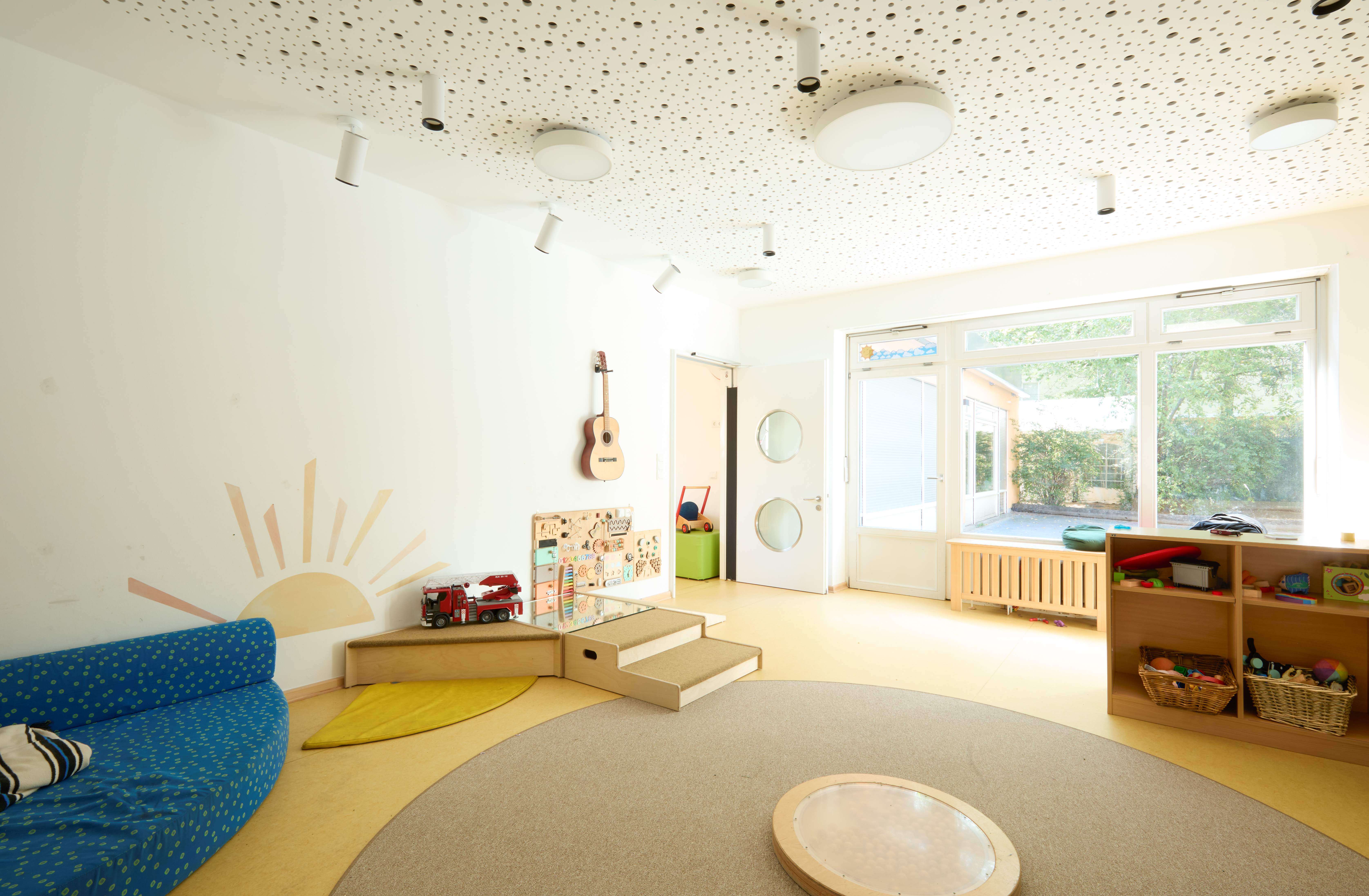 Heller Gruppenraum im Nestbereich mit großer Fensterfront und Tür zum Garten: links ein blaues Sofa, an der Wand Sonnenmalerei, eine hängende Gitarre und Spielwand; vorn runder Teppich, rechts Regale mit Körben und Spielzeug.