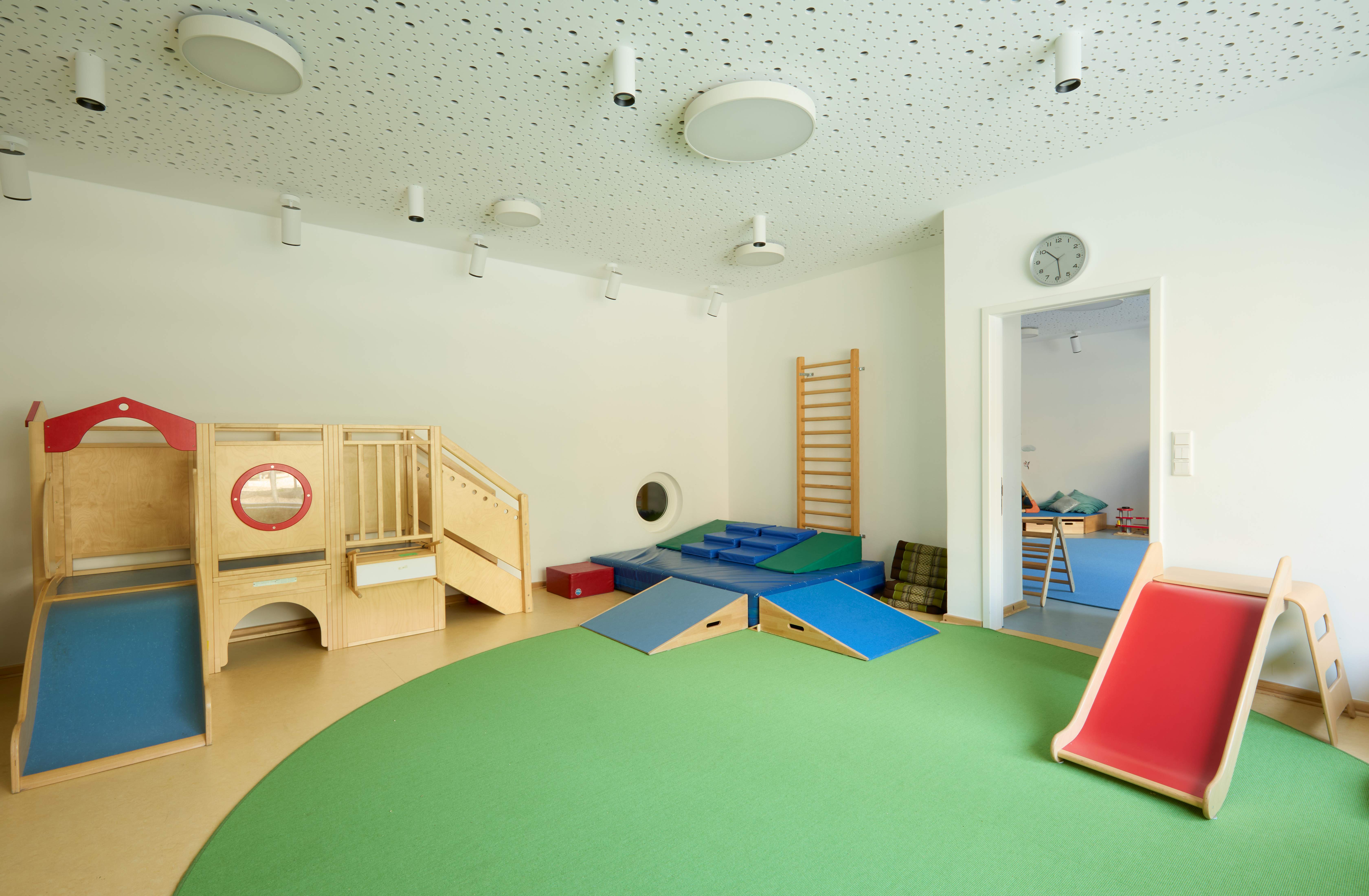 Heller Bewegungsraum im Nestbereich mit grünem Teppich: links ein kleines Holz‑Kletterhaus mit Rampe, rechts eine niedrige rote Rutsche; an der Wand Sprossenwand, daneben Tür zum angrenzenden Spielraum.