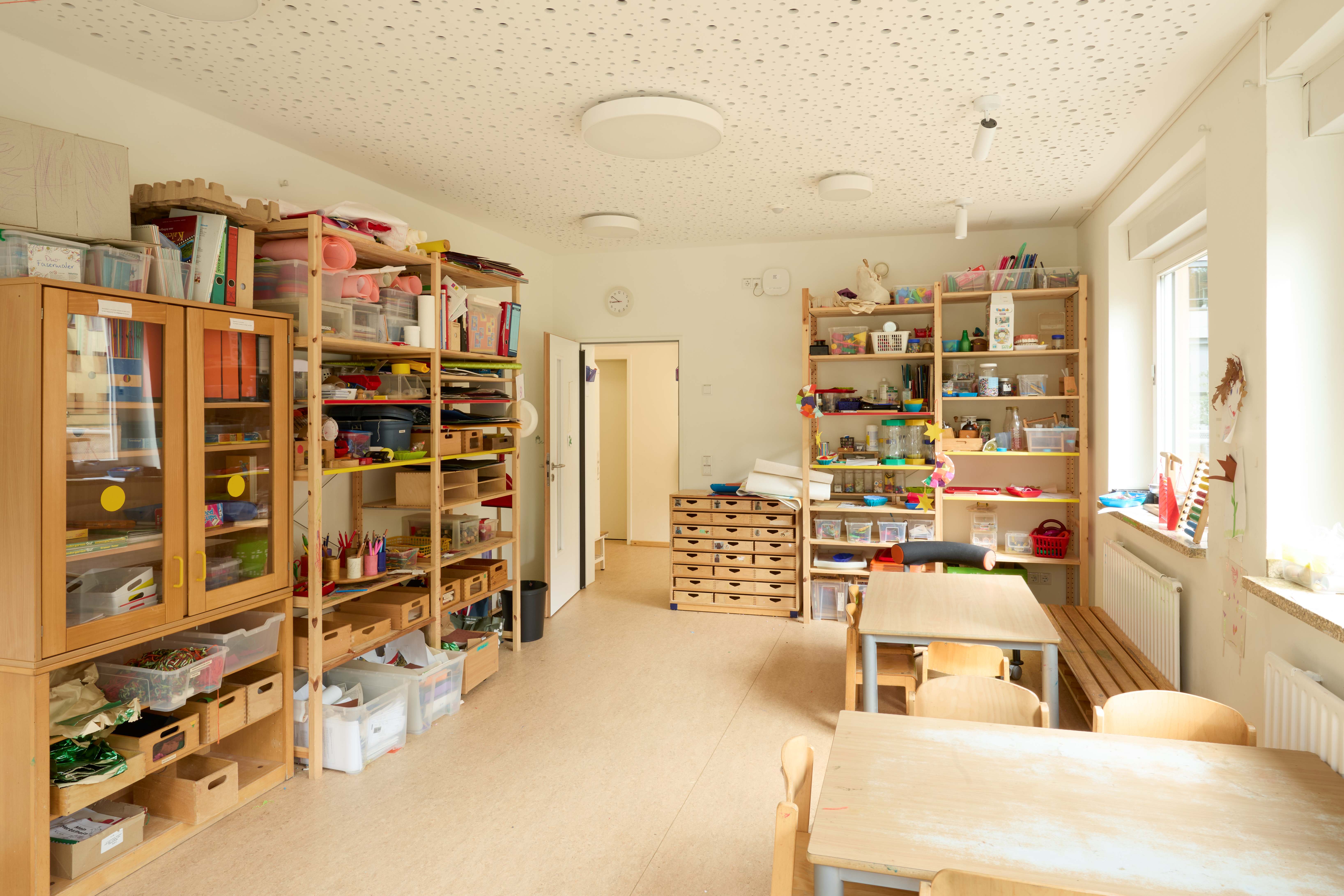 Heller Kreativ‑ und Werkraum in der Kita mit mehreren Holztischen und Kinderstühlen; rundum offene Regale und Schränke voller Bastelmaterialien.