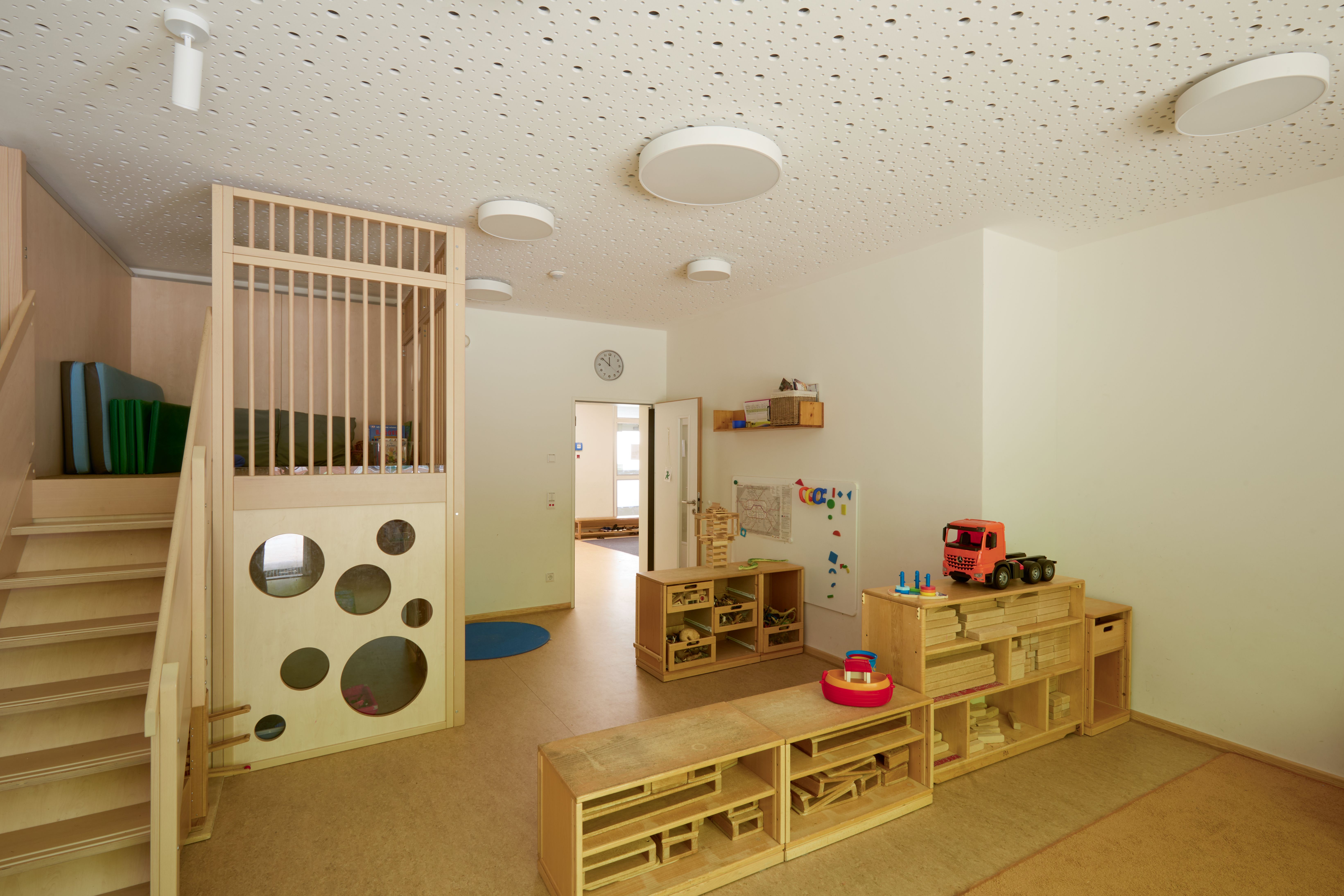 Heller Bauraum in der Kita: links führt eine kleine Treppe in eine erhöhte Spielebene; im Raum stehen niedrige Holzregale mit Bauklötzen und Spielzeugen.