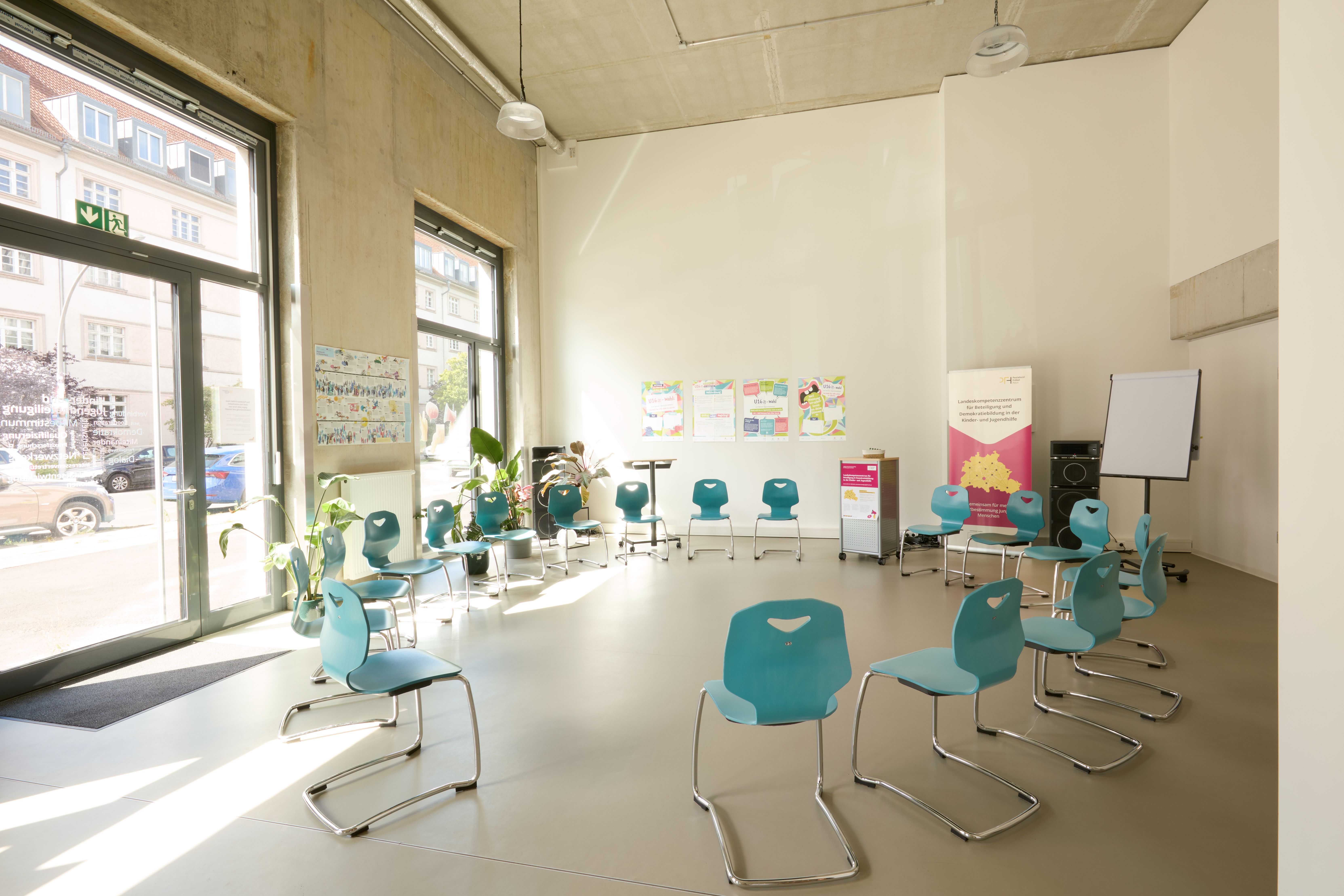 Heller Seminarraum mit großem Fenster zur Straße, türkisfarbene Stühle im Kreis angeordnet, grüne Topfpflanzen, Flipchart und Infoplakaten an der Wand.