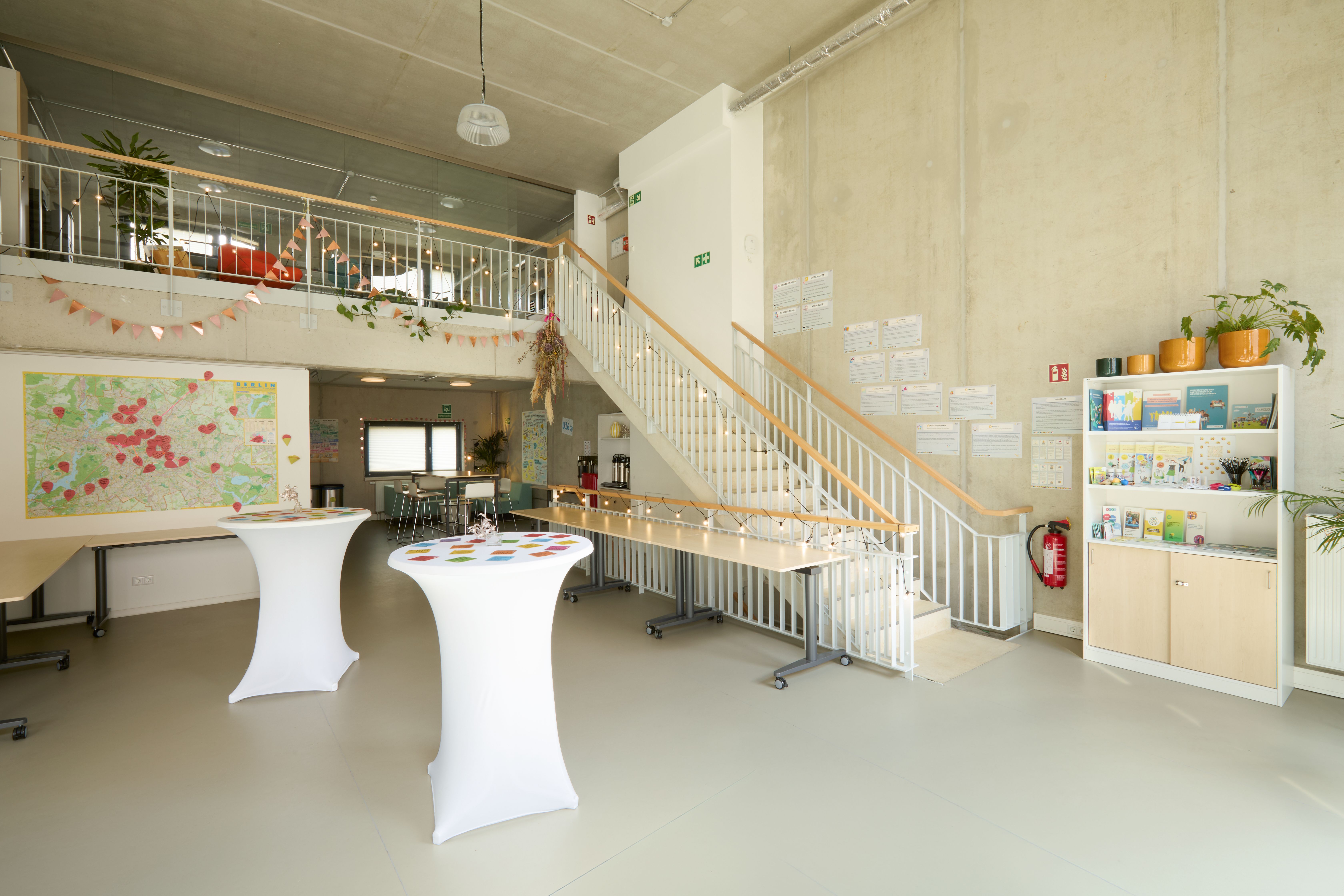 Großer heller Raum mit zwei weißen Stehtischen, Treppe mit Holzgeländer, Galerie oben mit Pflanzen, bunte Wimpelkette, Pinnwand mit roten Punkten, weiße Regale mit Büchern und Materialien rechts.