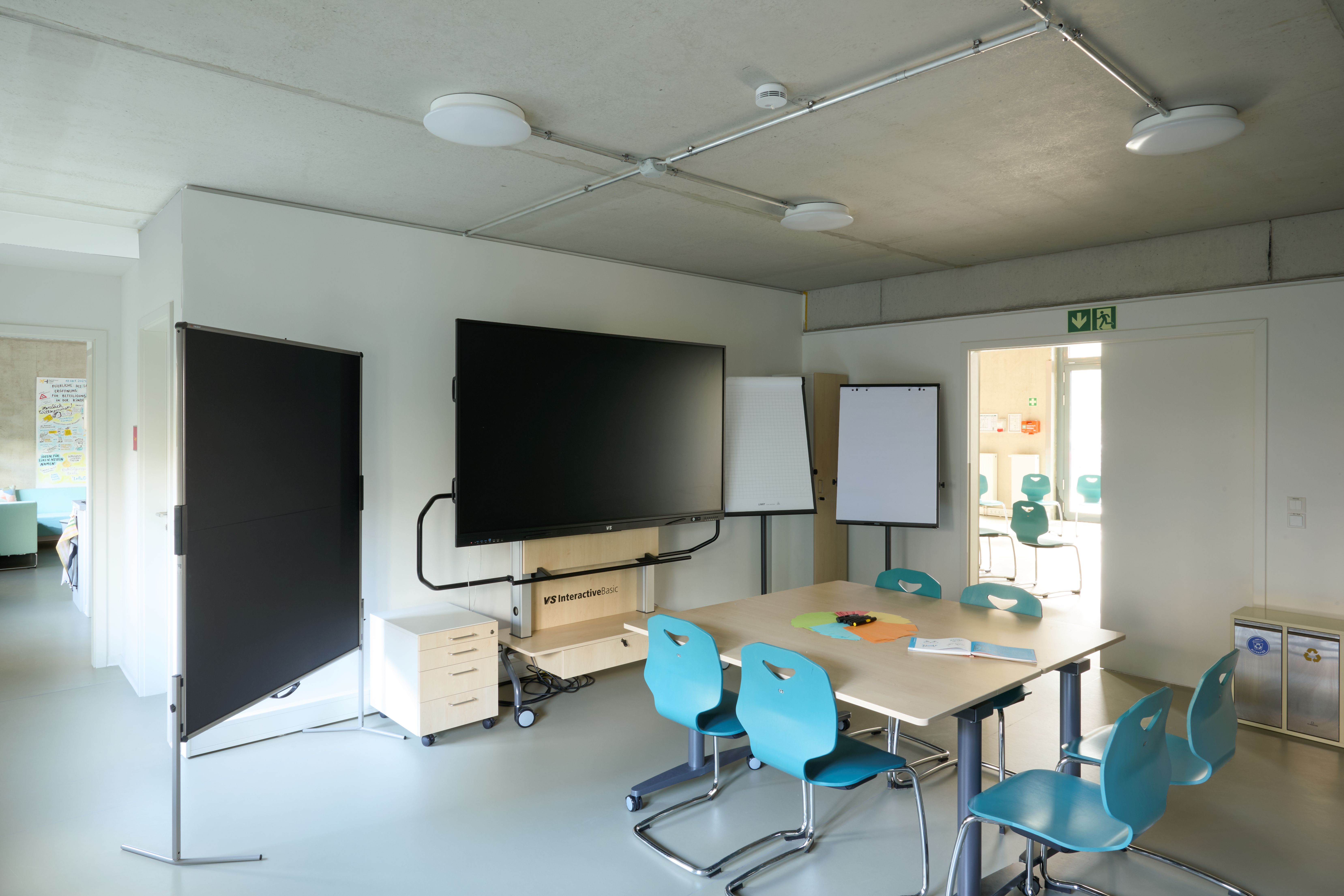 Schulungsraum mit Monitor, Steckwand und Whiteboards und beigem Tisch mit vier türkisfarbenen Stühlen.