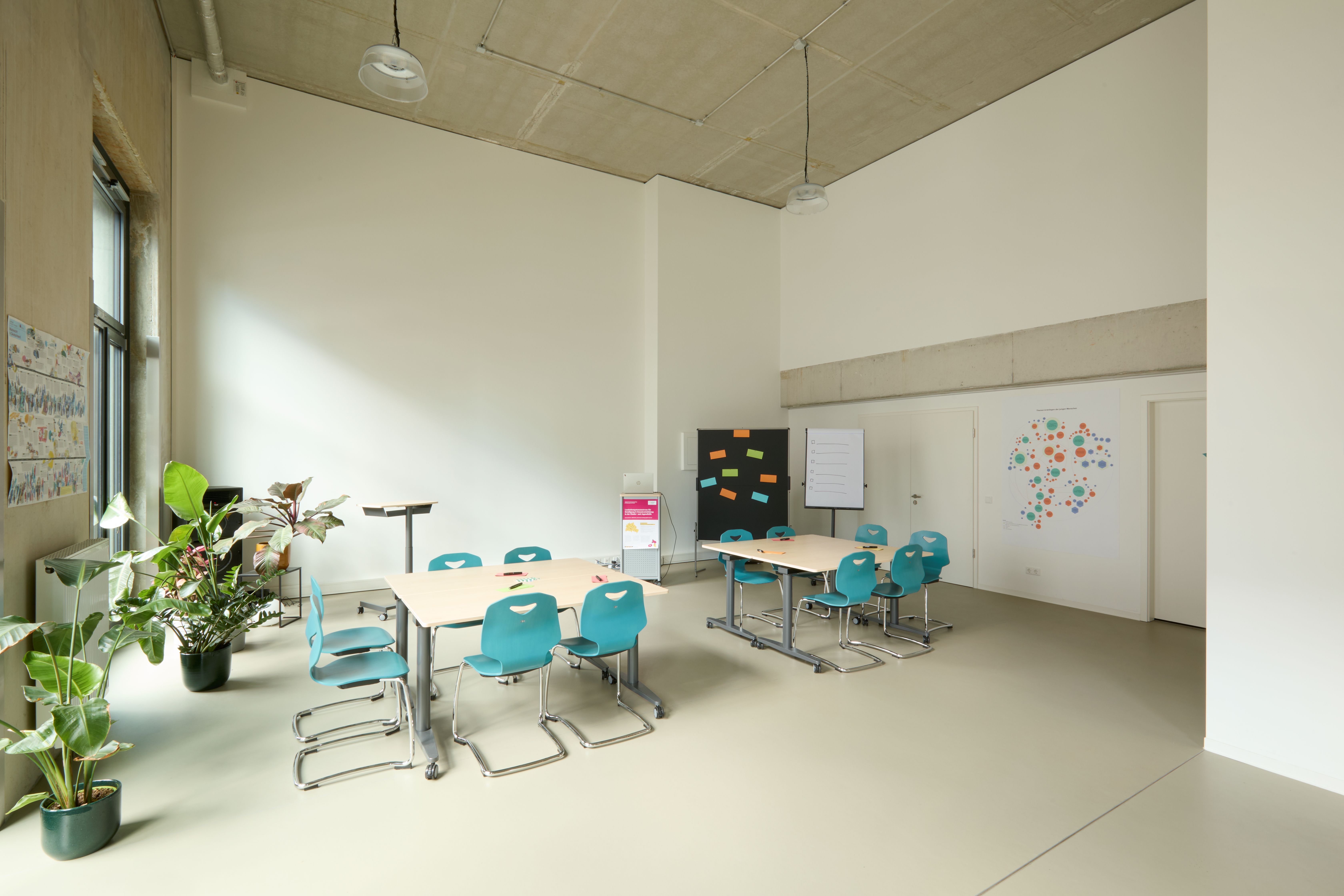 Großer heller Schulungsraum mit zwei beige Tischen, je vier türkisfarbenen Stühlen, großem Fenster mit Tageslicht. Grüne Pflanzen, Flipchart und Pinnwand mit bunten Punkten.