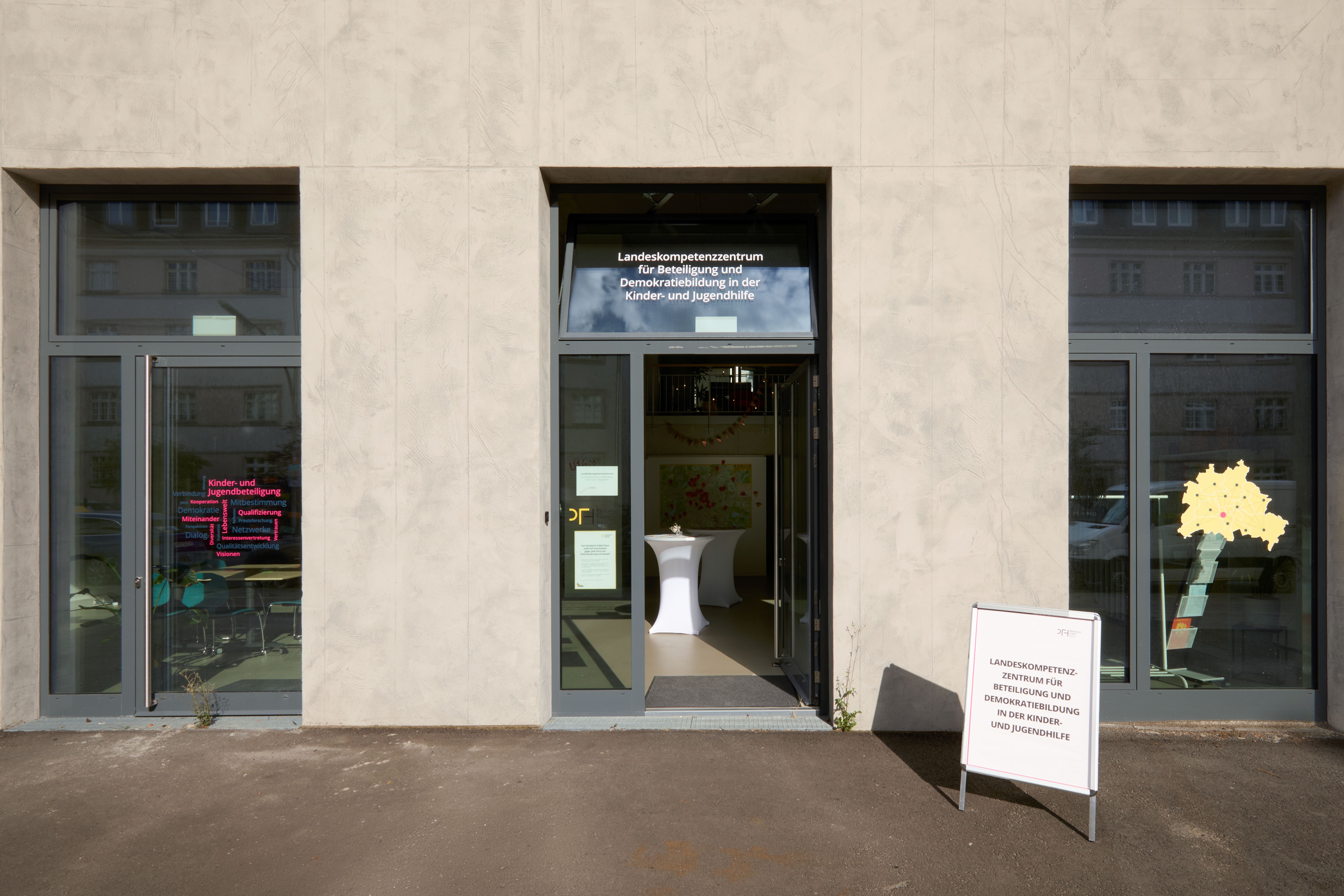 Moderne Gebäudefassade mit drei Glastüren. Mittlere Tür mit Beschilderung des Landeskompetenzzentrums. Linkes Fenster mit pinker Wortwolke, rechtes mit gelbem Umriss von Berlin. Vor der Tür steht ein Informationsständer.