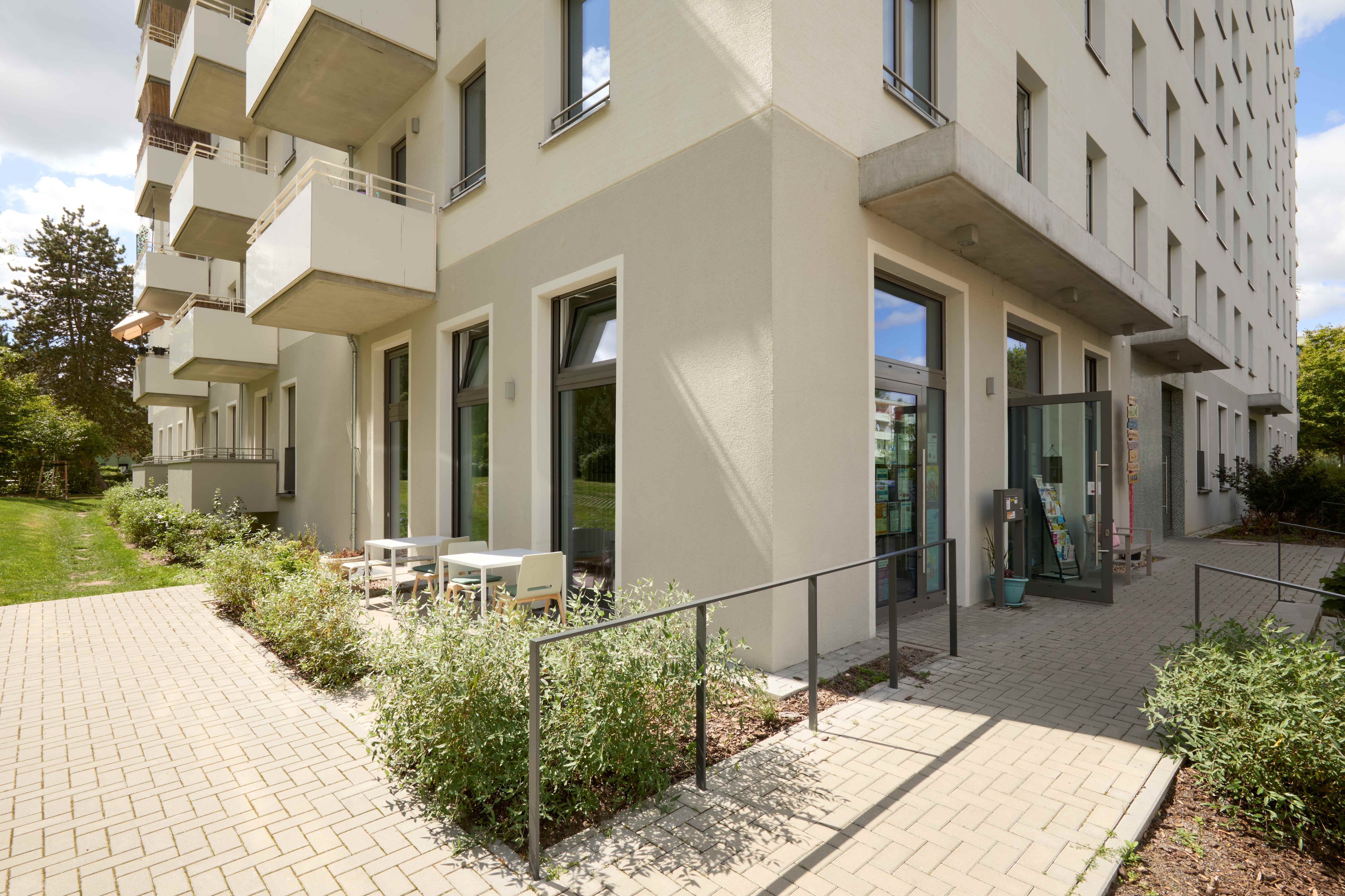 Außenansicht eines modernen, mehrstöckigen Gebäudes mit heller Fassade. Im Vordergrund gepflasterter Platz, grüne Hecken und Bepflanzung. Große Glastüren zum Eingangsbereich. Grüne Wiese und Bäume sichtbar. Sonniger Tag mit blauem Himmel.