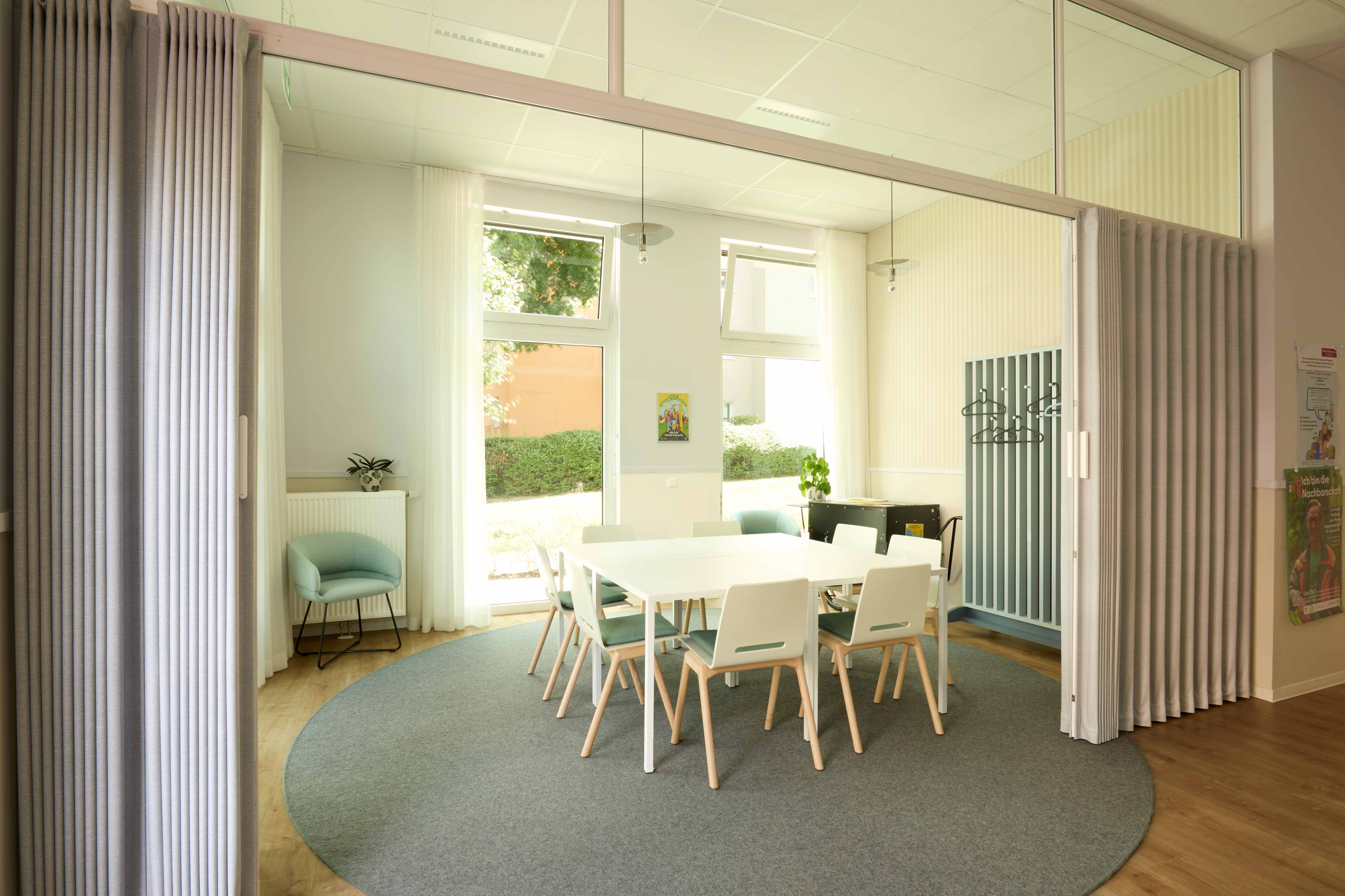 Heller Besprechungsraum mit rundem grauem Teppich, weißem Tisch, weißen Stühlen mit Holzbeinen, türkisem Sessel, großen Fenstern mit Gardinenschienen und grünen Pflanzen. Moderne, offene Raumgestaltung mit Tageslicht.