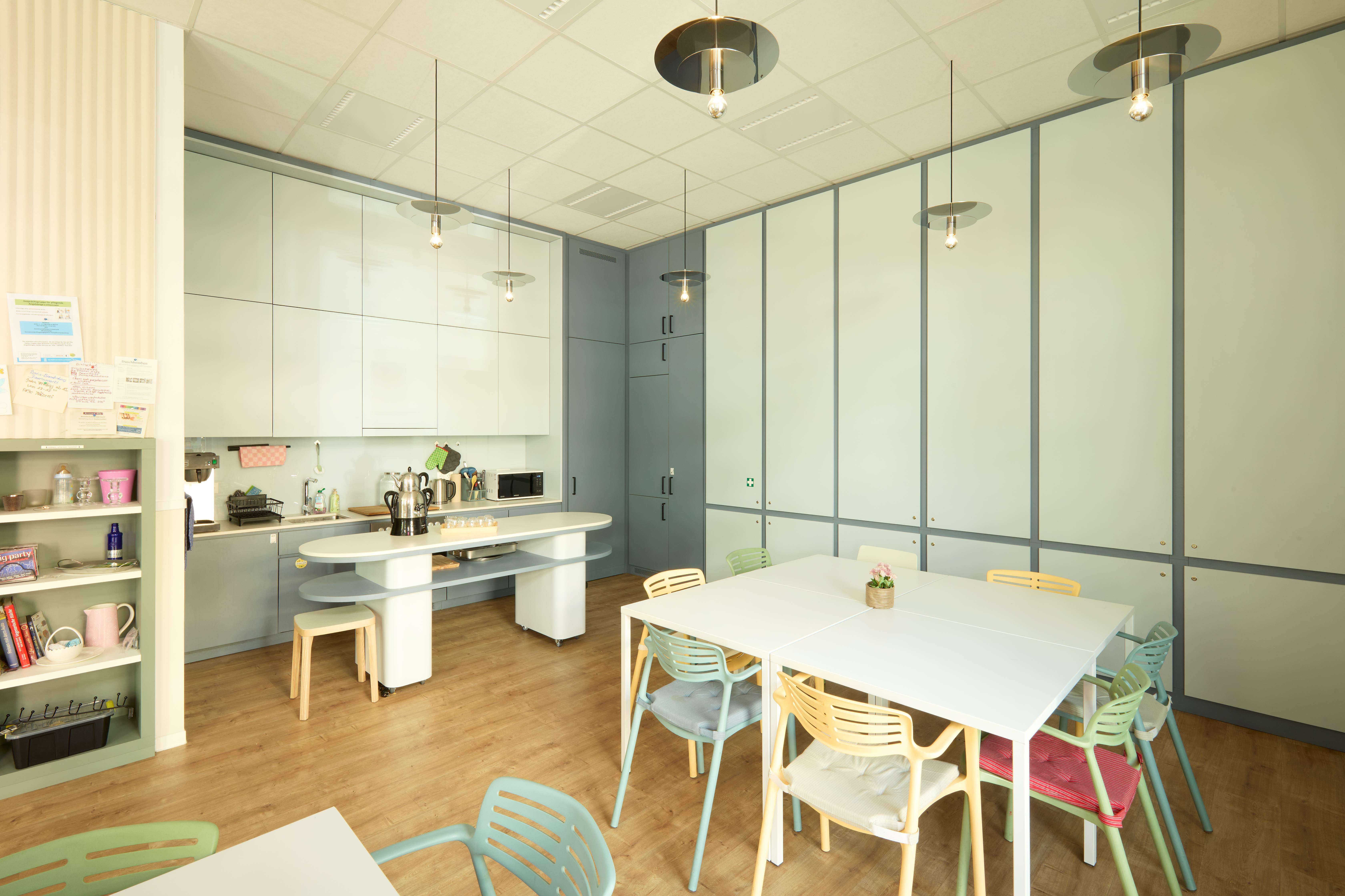 Moderne Gemeinschaftsküche mit weißen Oberschränken, graugrünen Unterschränken, Kochinsel mit weißer Arbeitsplatte und Holzhocker. Großer weißer Esstisch mit türkis-grünen Stühlen. Regale mit Küchenutensilien und Dekoration. Heller Raum mit Holzboden.
