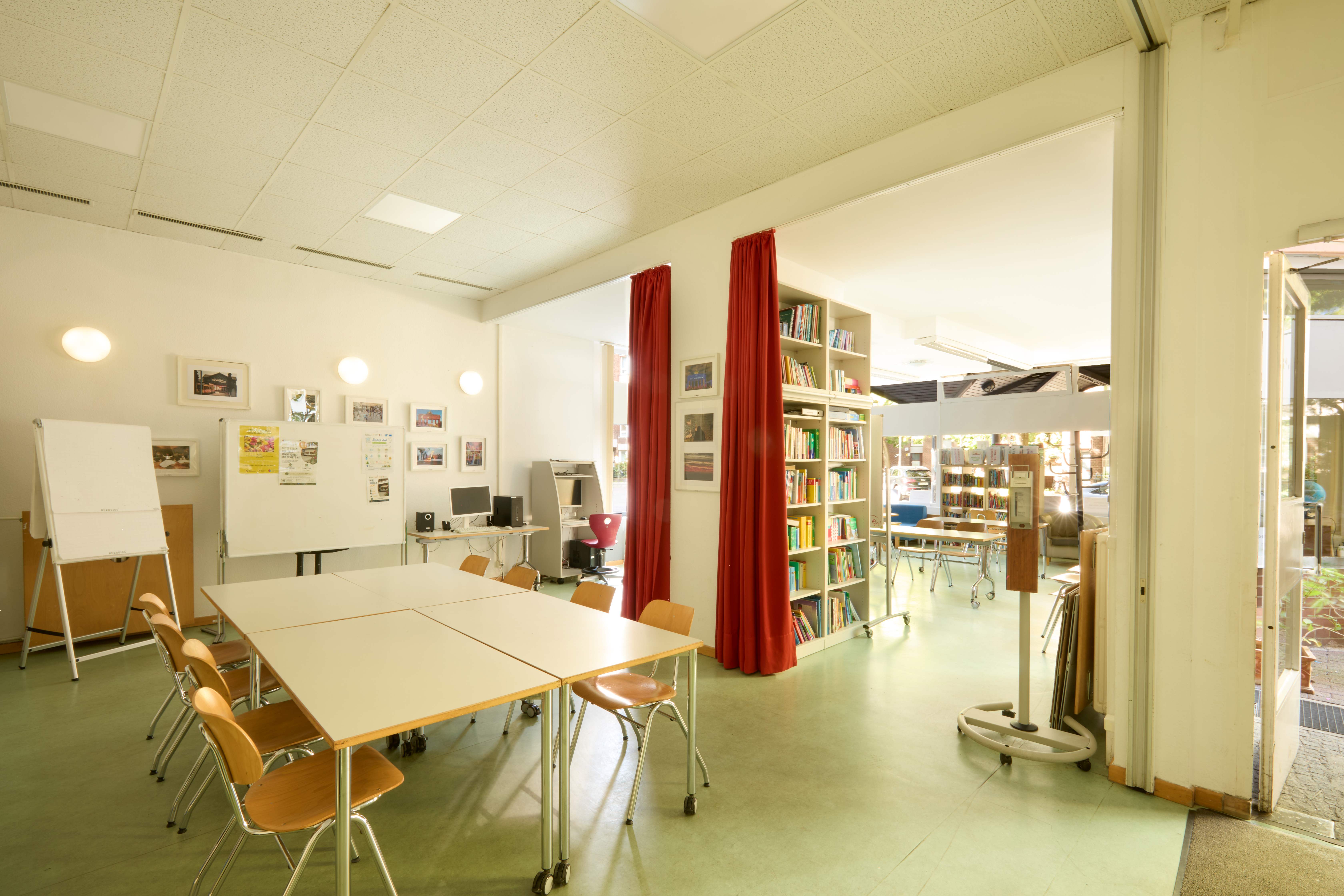 Offener Gruppenbereich mit zusammengestellten Tischen, Stühlen und Whiteboard. In der Raummitte hohe Bücherregale, rote Vorhänge als Raumteiler und Computerarbeitsplätze im Hintergrund.