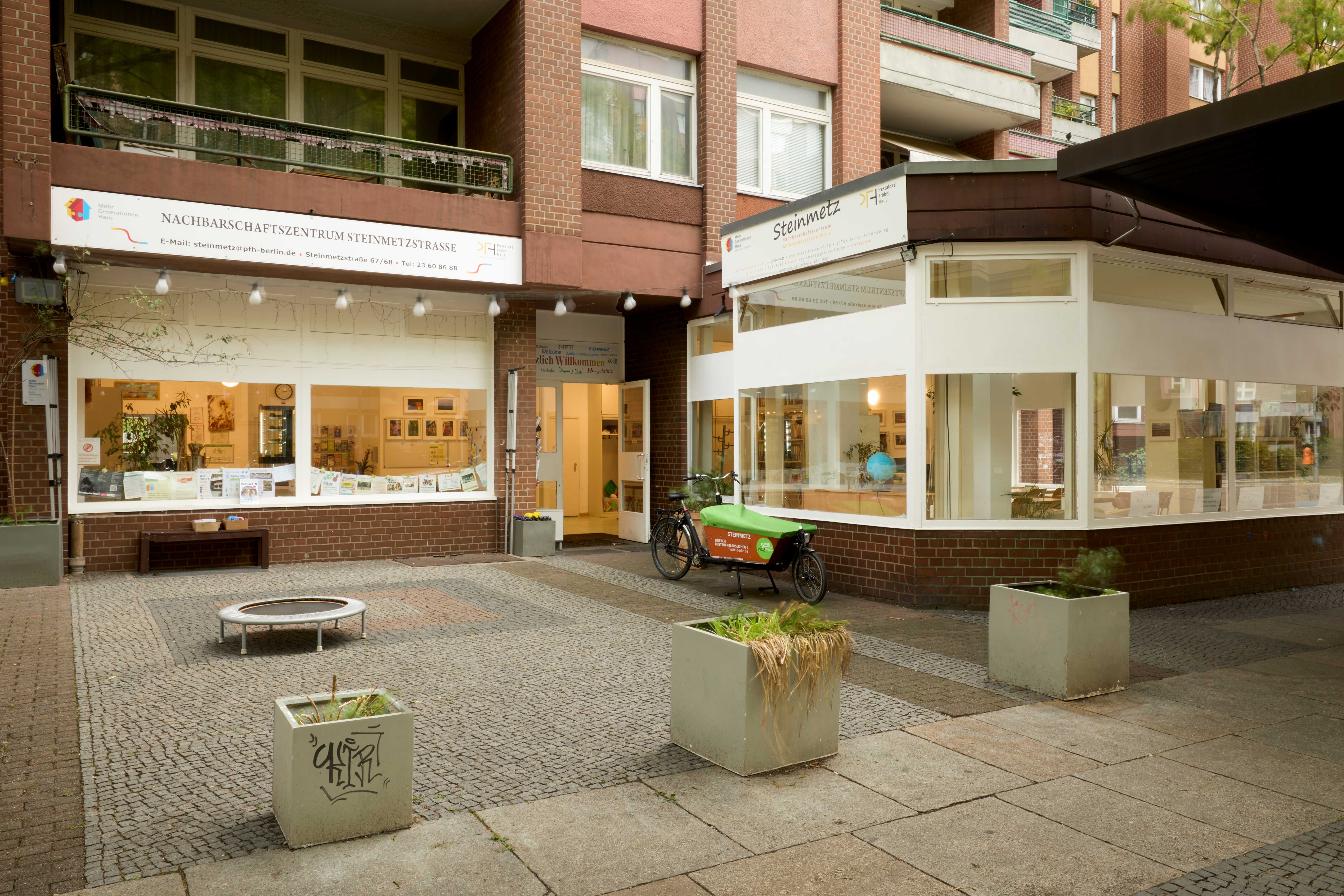  Außenansicht des Nachbarschaftszentrums in einem roten Klinkerbau. Große Schaufenster geben den Blick in helle Räume frei; vor dem Eckfenster steht ein grünes Lastenrad. Davor ein kleiner gepflasterter Platz mit Pflanzkübeln.