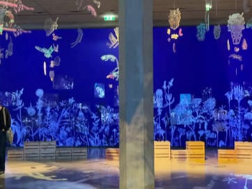 Innenansicht eines Ausstellungsraums mit einer blauen Wand im Hintergrund, die mit floralen und tierischen Motiven dekoriert ist. Hängende, transparente Kunstobjekte in verschiedenen Farben, darunter rosa und gelb, sind von der Decke abgehängt. In der Mitte des Raumes steht ein Säulenpfeiler, und der Boden ist mit Schatten und Lichtmustern versehen. Eine Person ist im Hintergrund zu sehen, die den Raum betrachtet.