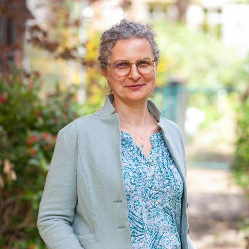 Portrait von Wioletta Wendland: Eine Frau trägt eine hellblaue Bluse und Strickjacke sowie eine Brille. Sie lächelt. Sie steht draußen vor grünen Bäumen und lächelt. Der Hintergrund ist verschwommen.